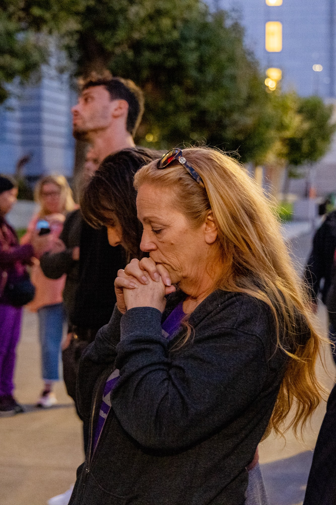 A crowd of people stands somberly outdoors, with one woman in the foreground clasping her hands in a prayerful gesture, surrounded by trees and buildings.