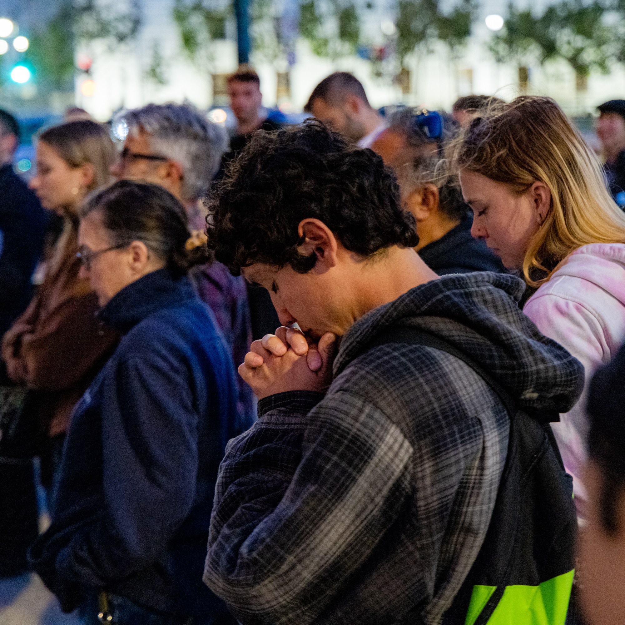 A group of people are standing close together, appearing solemn. Many have their heads bowed and eyes closed, possibly in prayer or reflection.