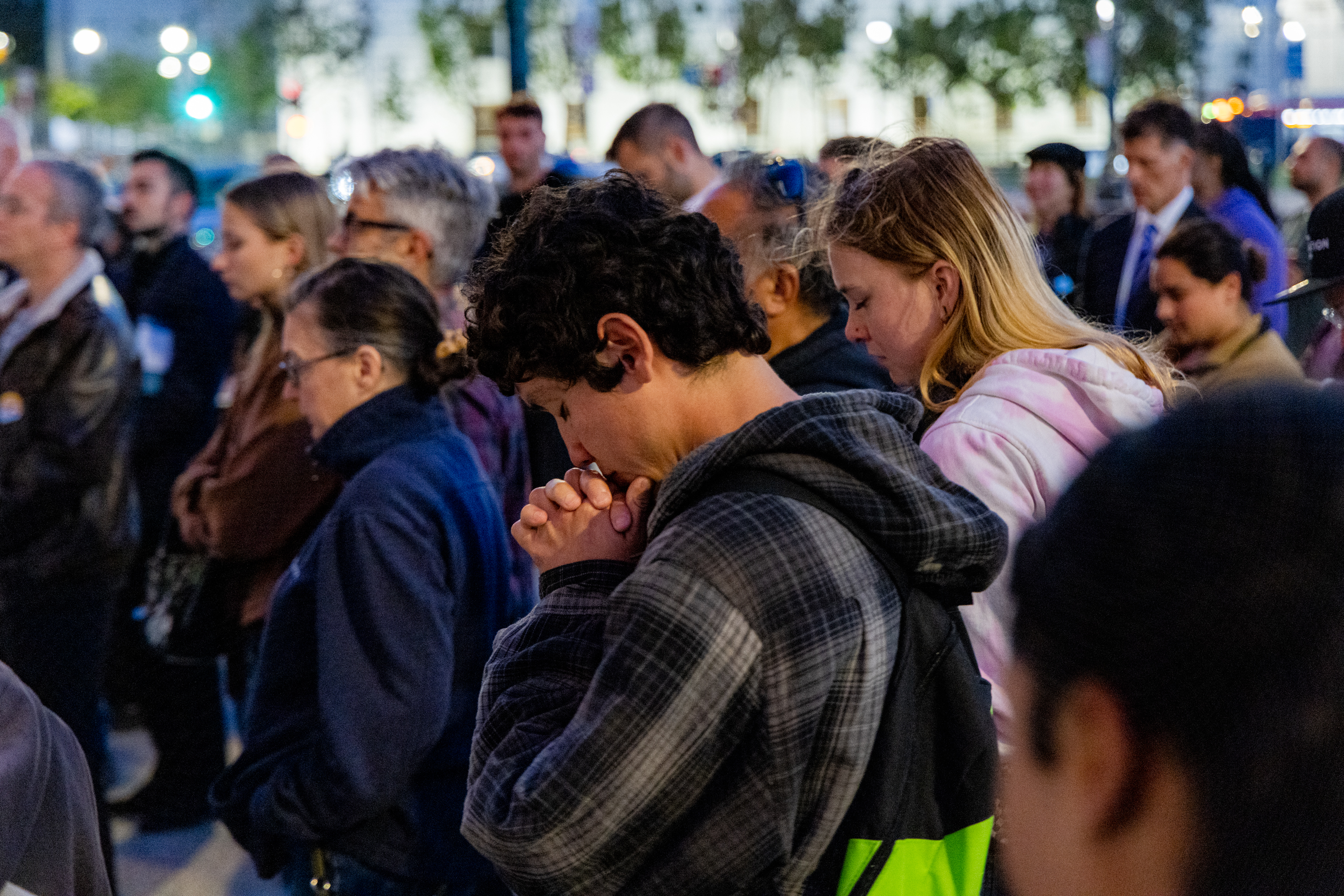 A group of people are standing close together, appearing solemn. Many have their heads bowed and eyes closed, possibly in prayer or reflection.