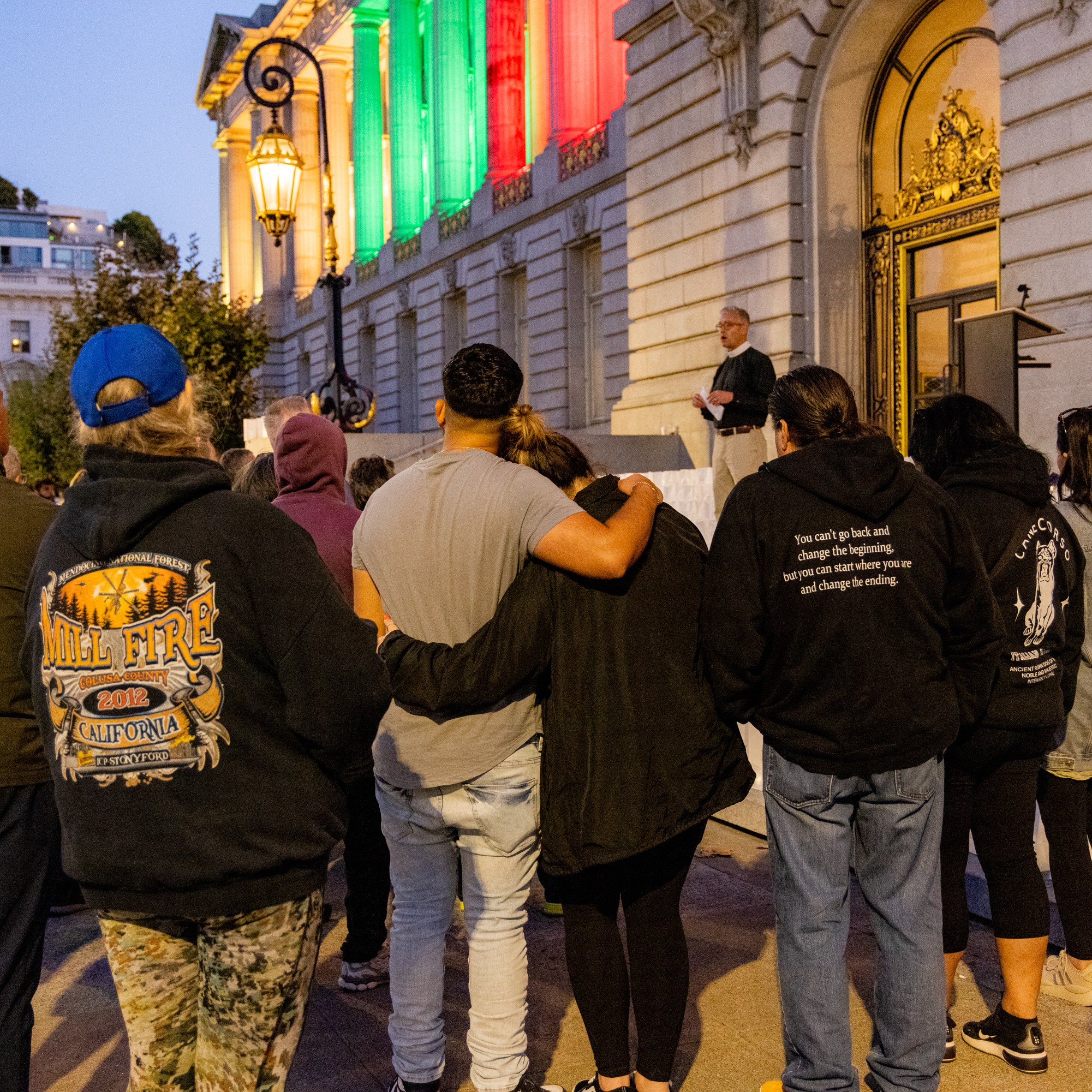 A group of people gather outside a building lit with green and red lights, watching a speaker. Some individuals are hugging, creating a sense of community.