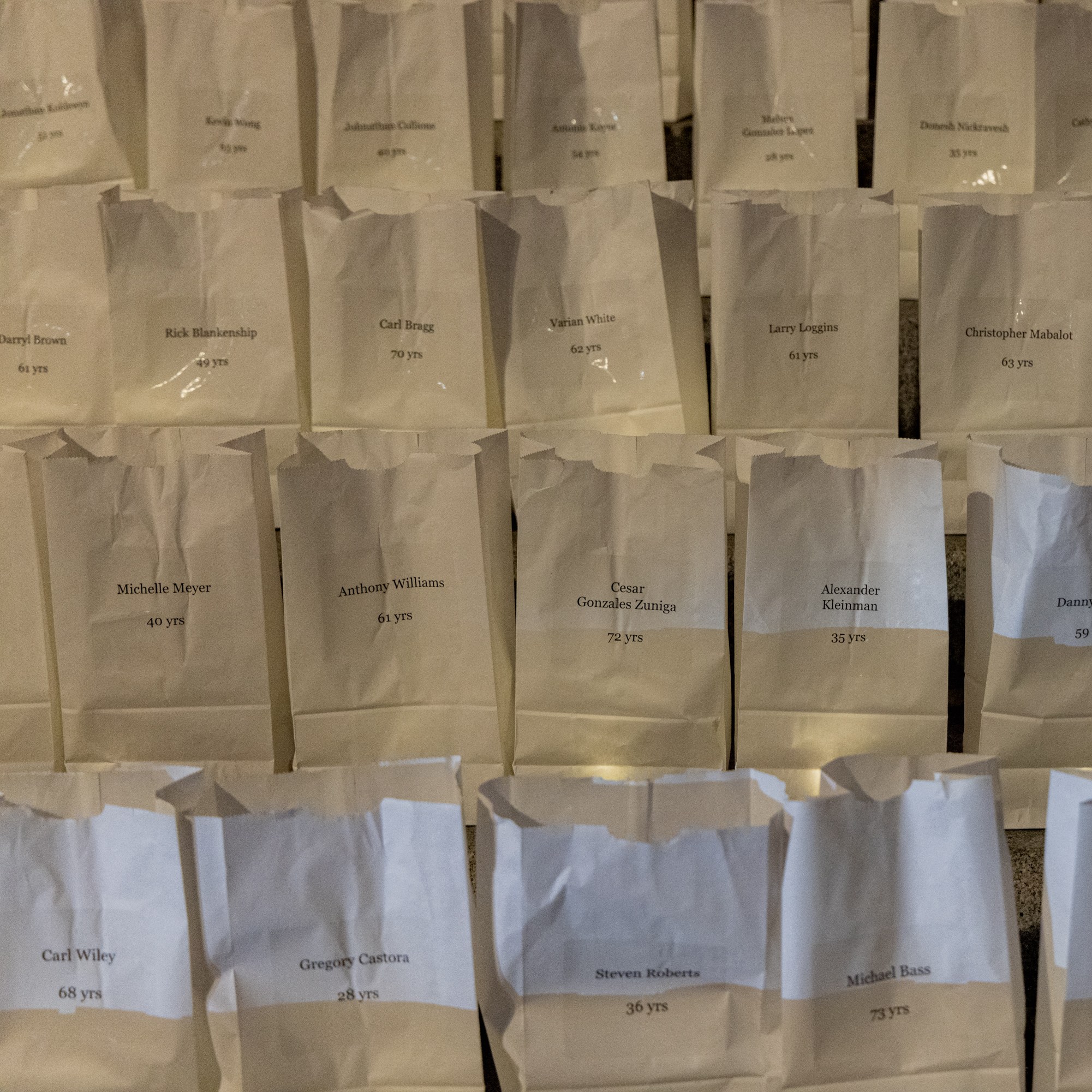 The image shows rows of white paper bags, each labeled with a name and age, arranged in a grid-like pattern. The bags appear to be part of a display or memorial.