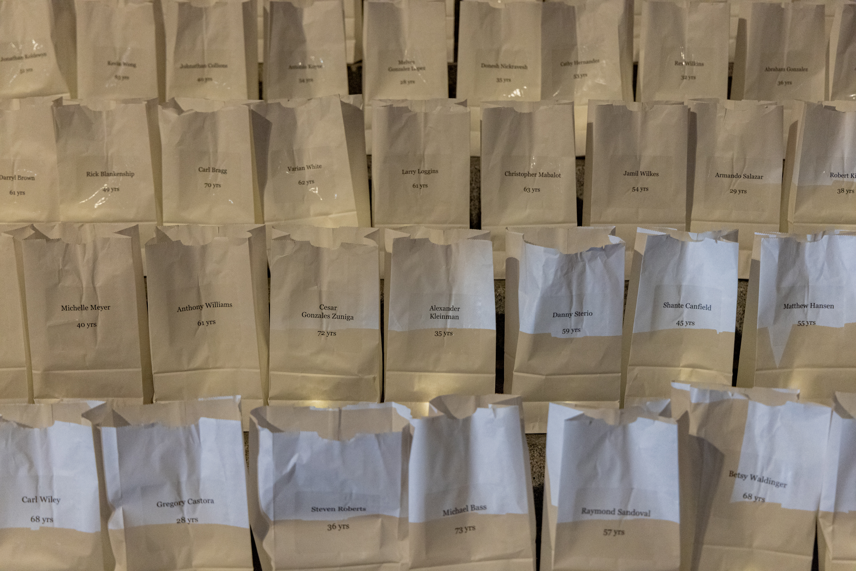 The image shows rows of white paper bags, each labeled with a name and age, arranged in a grid-like pattern. The bags appear to be part of a display or memorial.