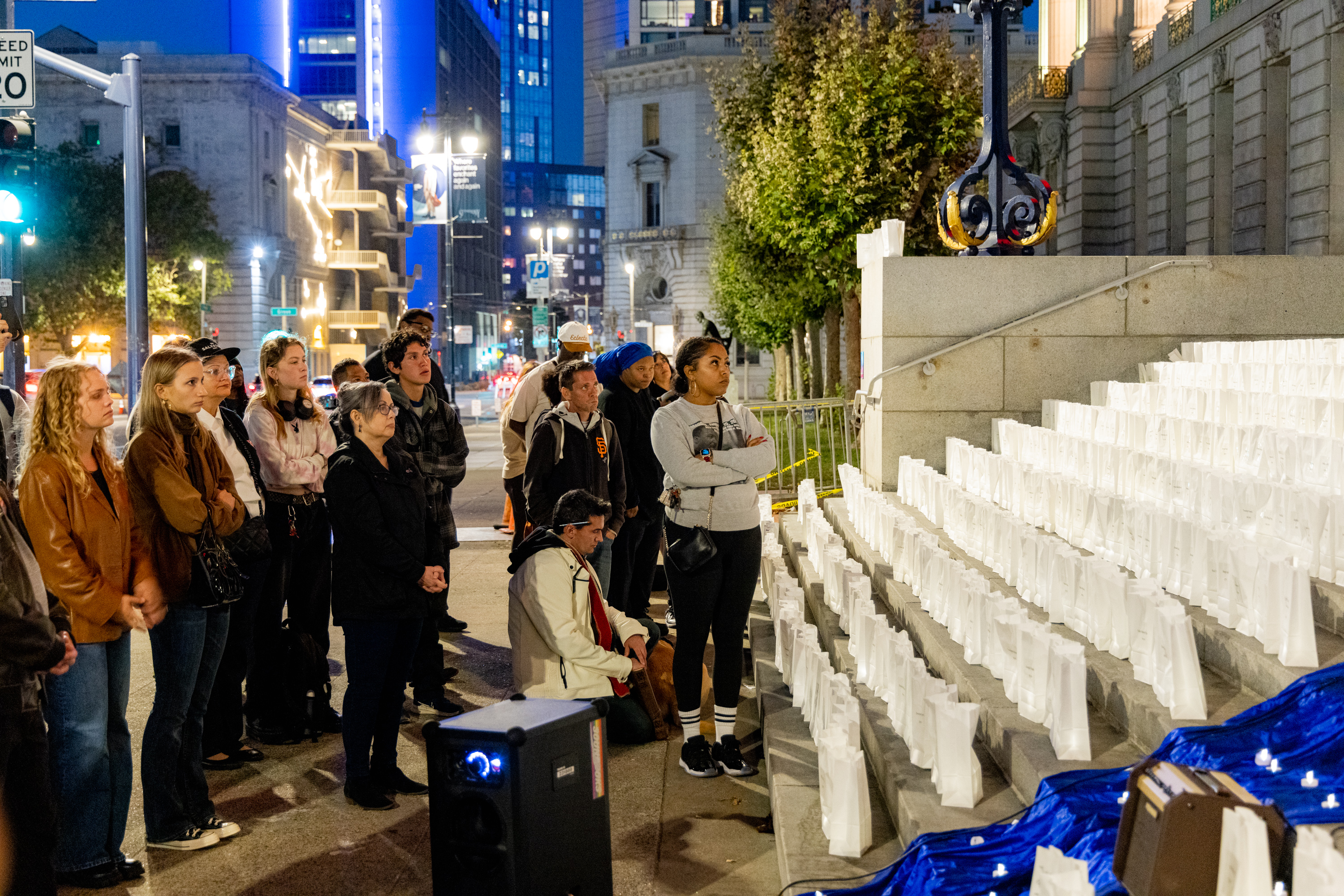 A group of people stands solemnly in an urban setting at night, facing a staircase lined with white paper lanterns. The city lights and buildings illuminate the background.