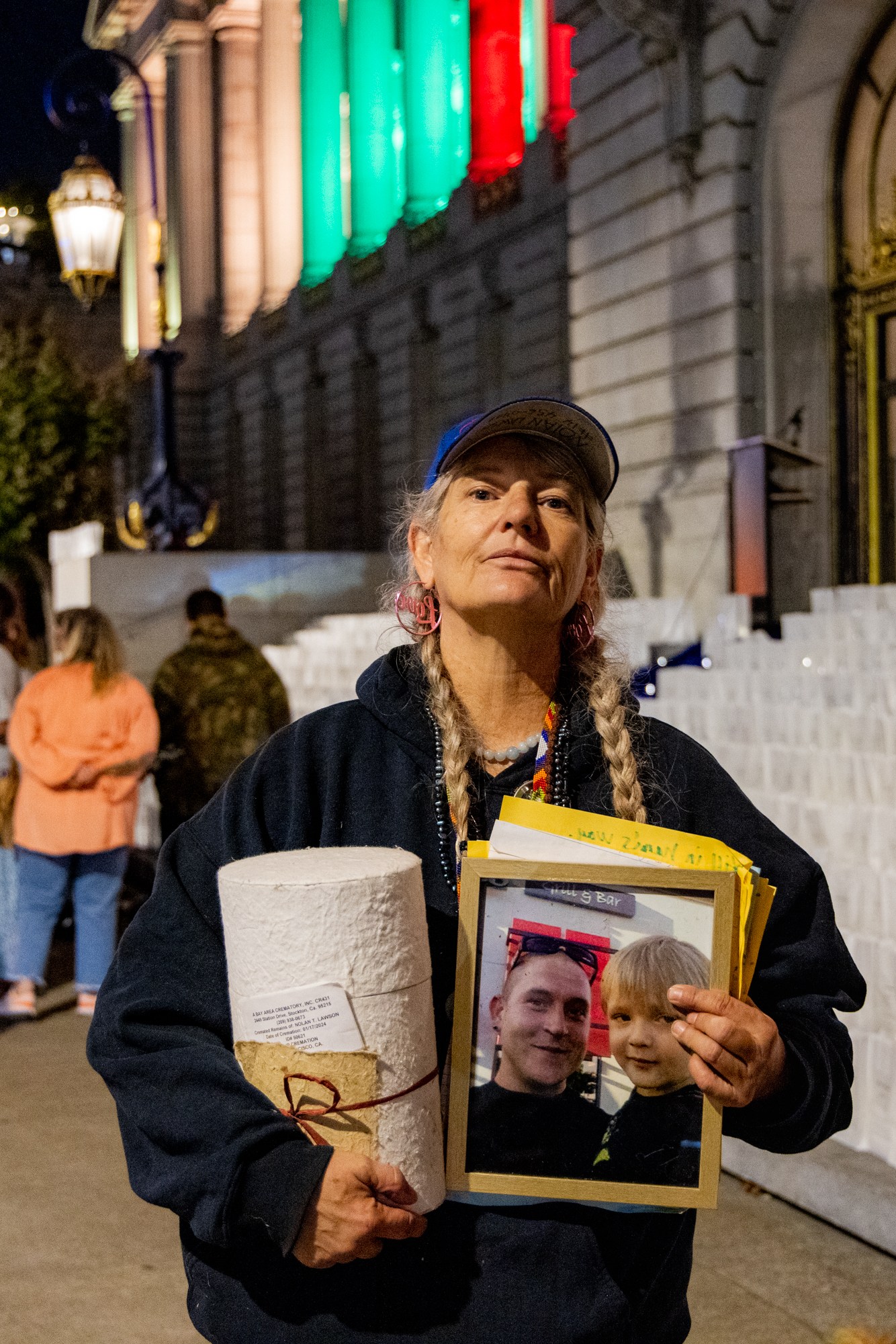 A woman stands in front of a lit-up building at night, holding a photo of a man and a child, and a rolled-up paper. People and lanterns can be seen in the background.