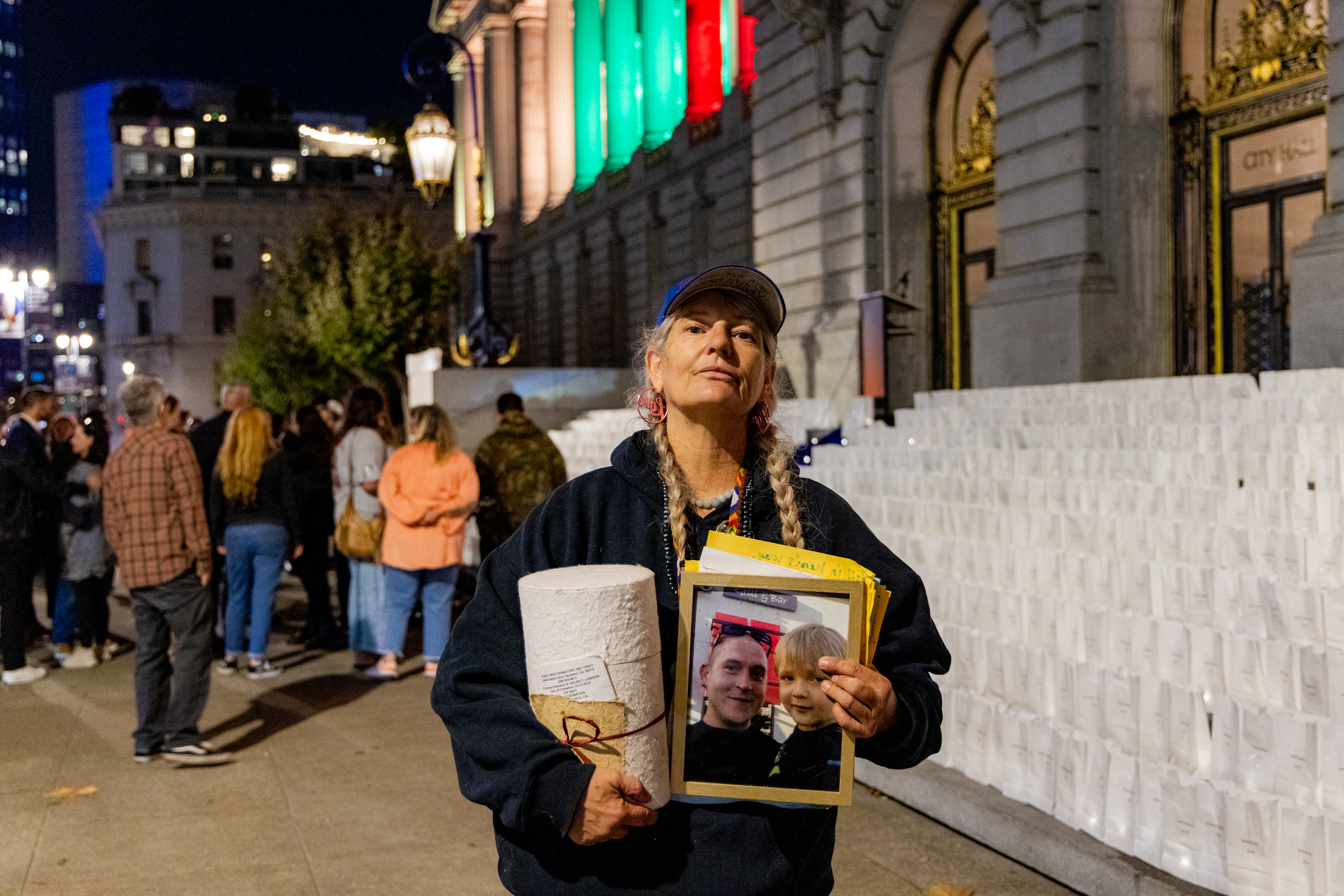 A woman stands in front of a lit-up building at night, holding a photo of a man and a child, and a rolled-up paper. People and lanterns can be seen in the background.