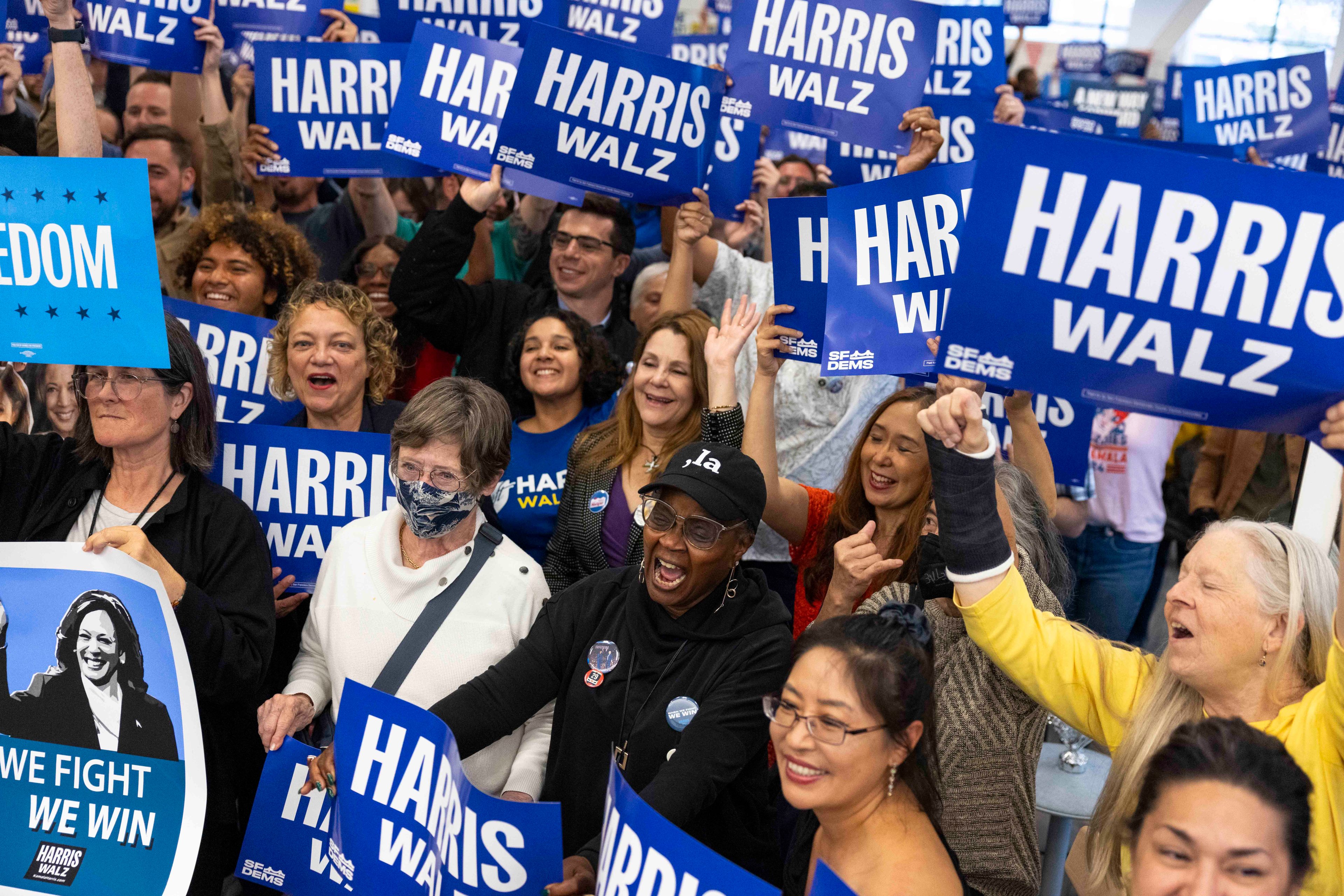 A joyous, diverse crowd holds "Harris Walz" signs, some yelling, promoting unity and support at what appears to be a political rally.