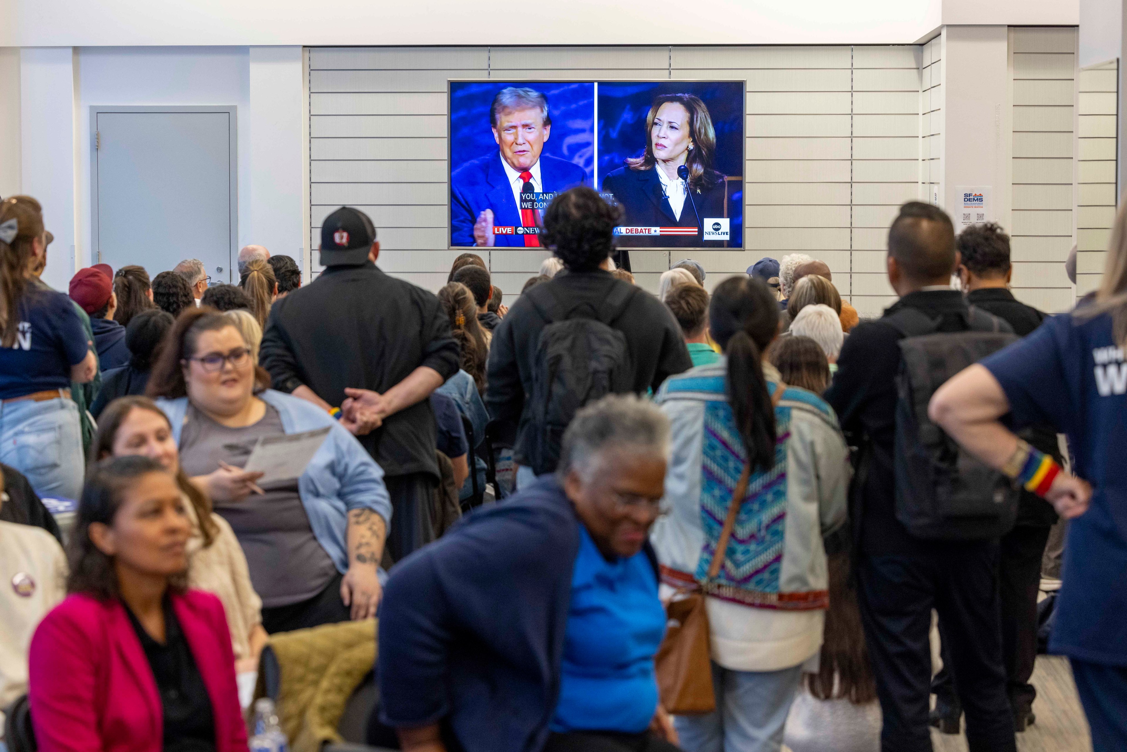 A crowd watches a debate on a wall-mounted TV in a large room, featuring two political figures. The audience is diverse, standing and seated, focused on the screen.