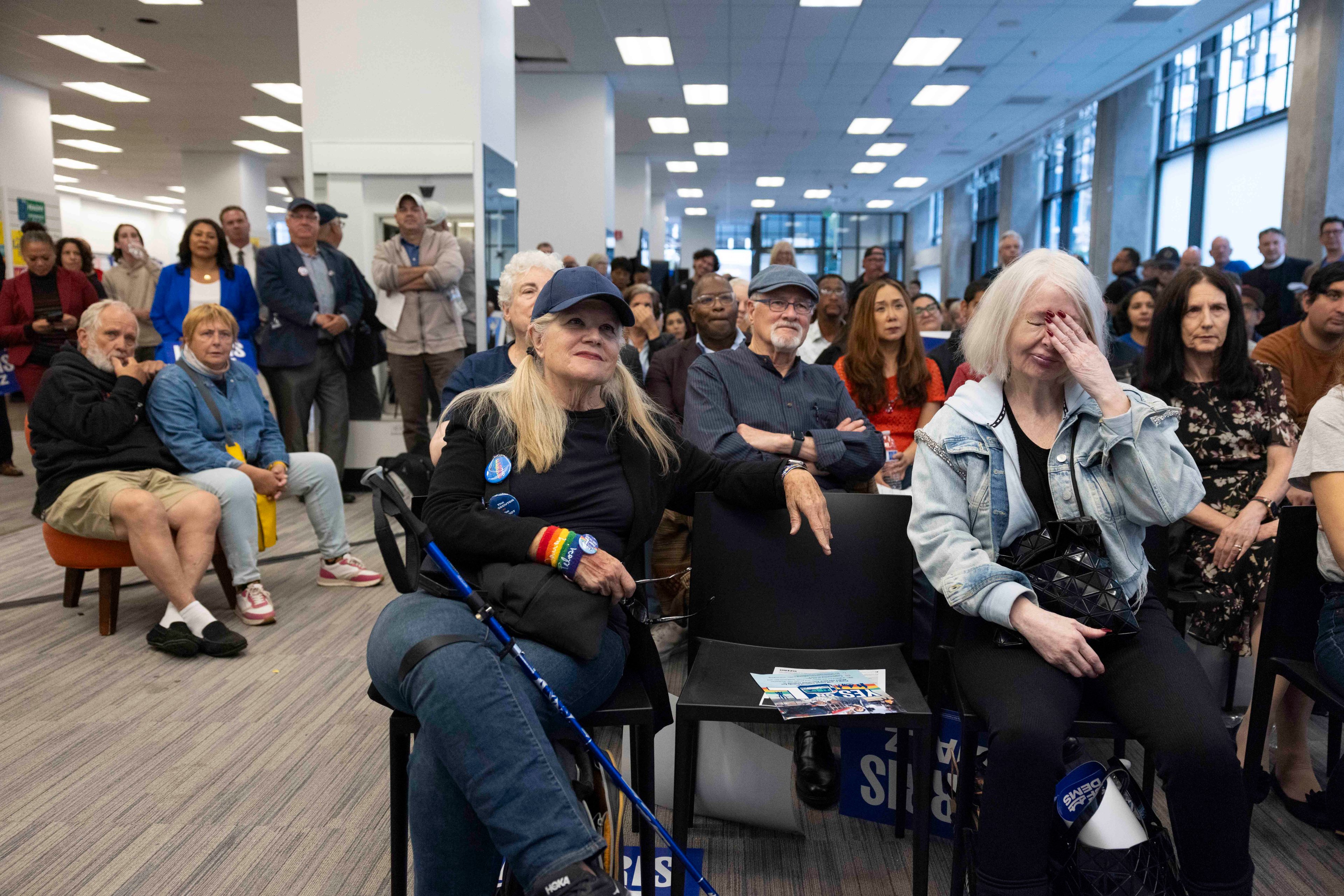 A diverse group of people sits attentively in a spacious, brightly lit room, with some standing in the back. Two women in the foreground appear emotional; one wipes her eye.