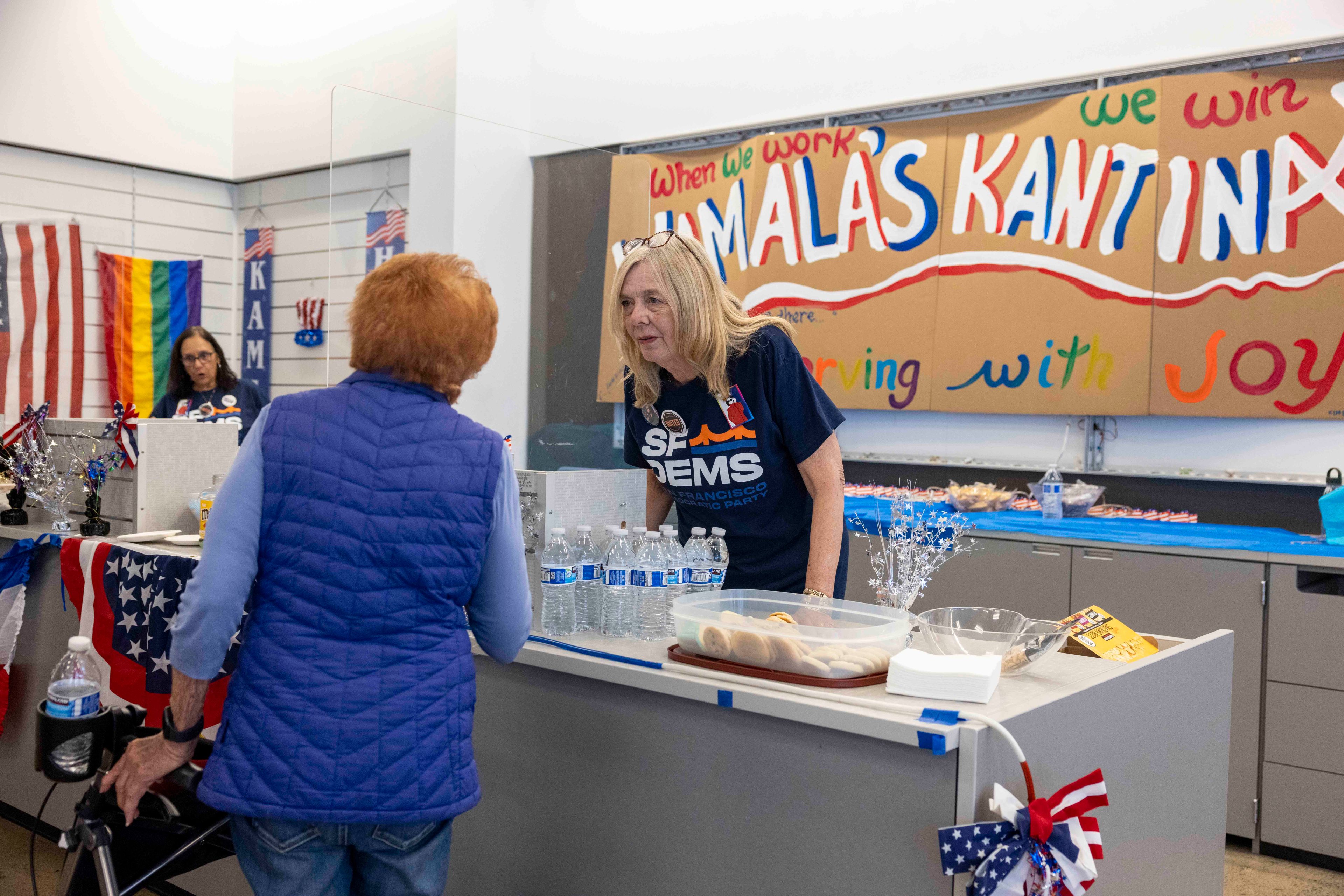 Two women are at a decorated counter with snacks and drinks, including bottled water. One wears a &quot;SF DEMS&quot; shirt, and a sign reads &quot;Kamala's Kantina,&quot; with colorful banners.