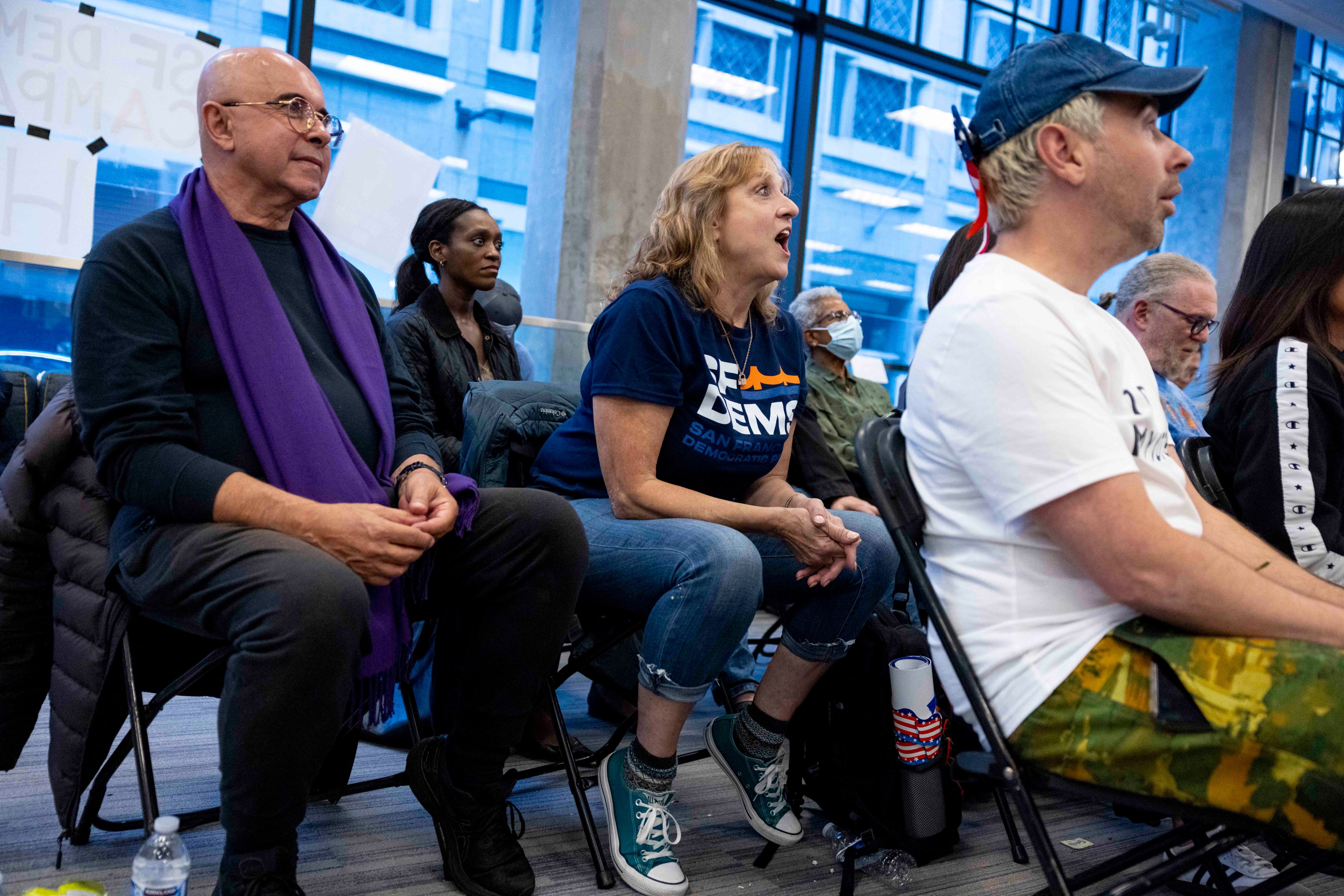 A group of people sits attentively in a room with large windows. One person wears a &quot;Dems&quot; t-shirt and another sports a purple scarf. Some wear masks, and all are focused.