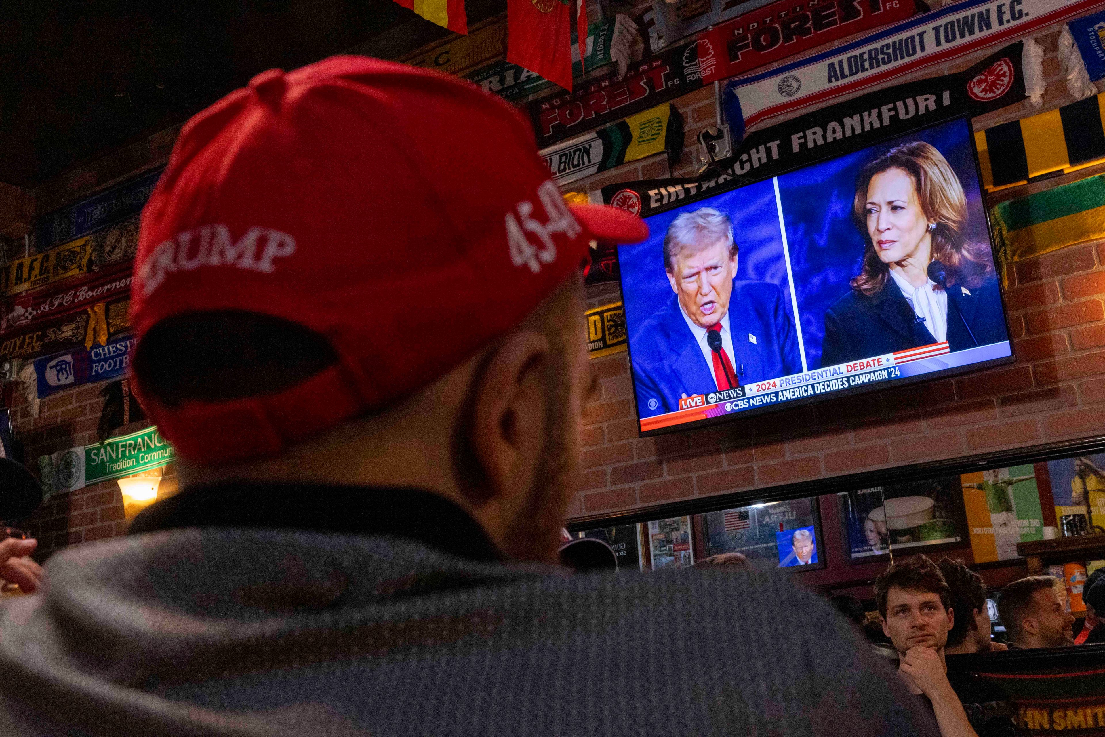 A person wearing a red &quot;Trump 45&quot; cap watches a televised debate between two politicians in a brick-walled room decorated with numerous sports team scarves.