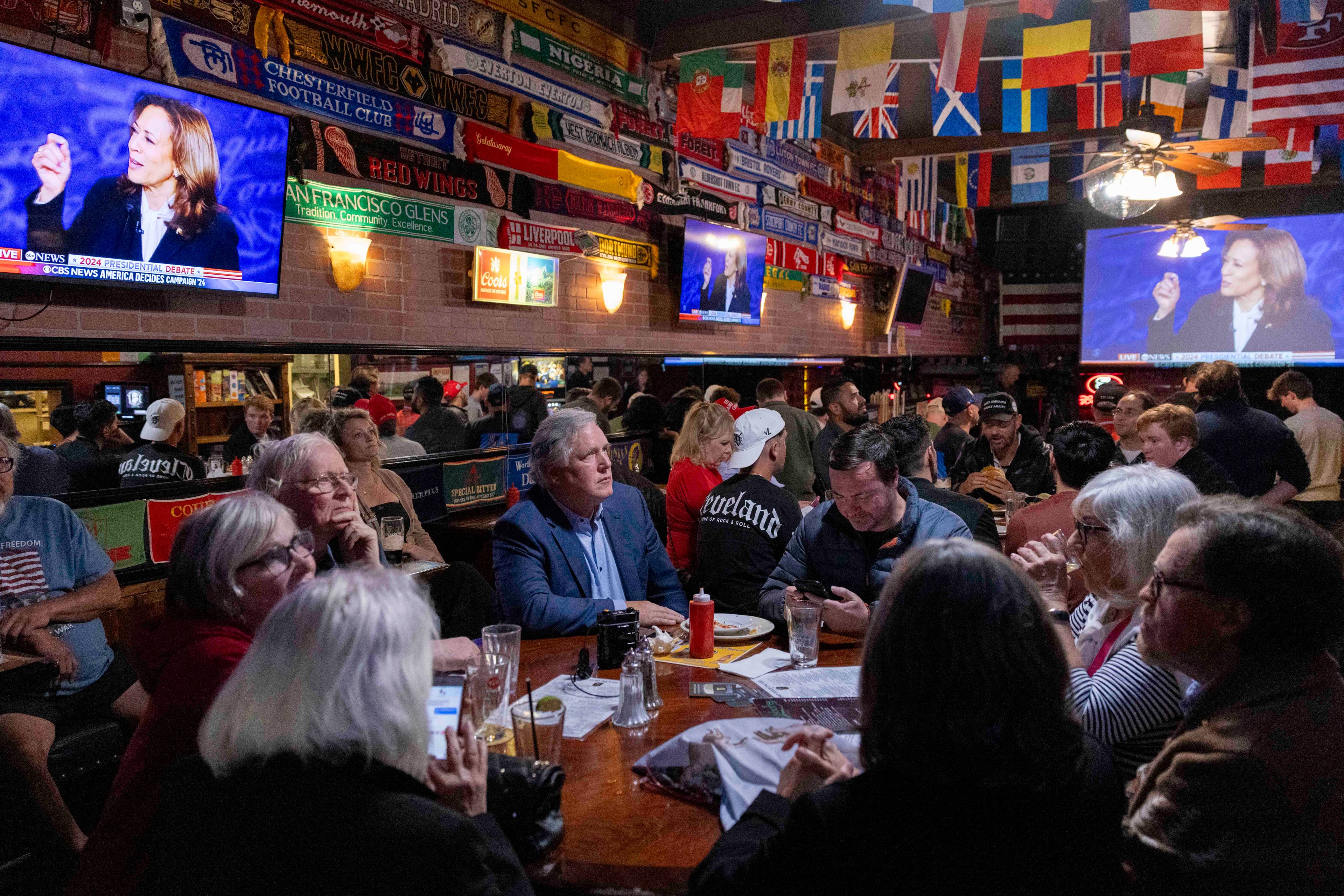 A group of people in a pub is watching a televised debate. Flags and sports scarves adorn the walls and multiple TVs show the same debate, creating a lively atmosphere.