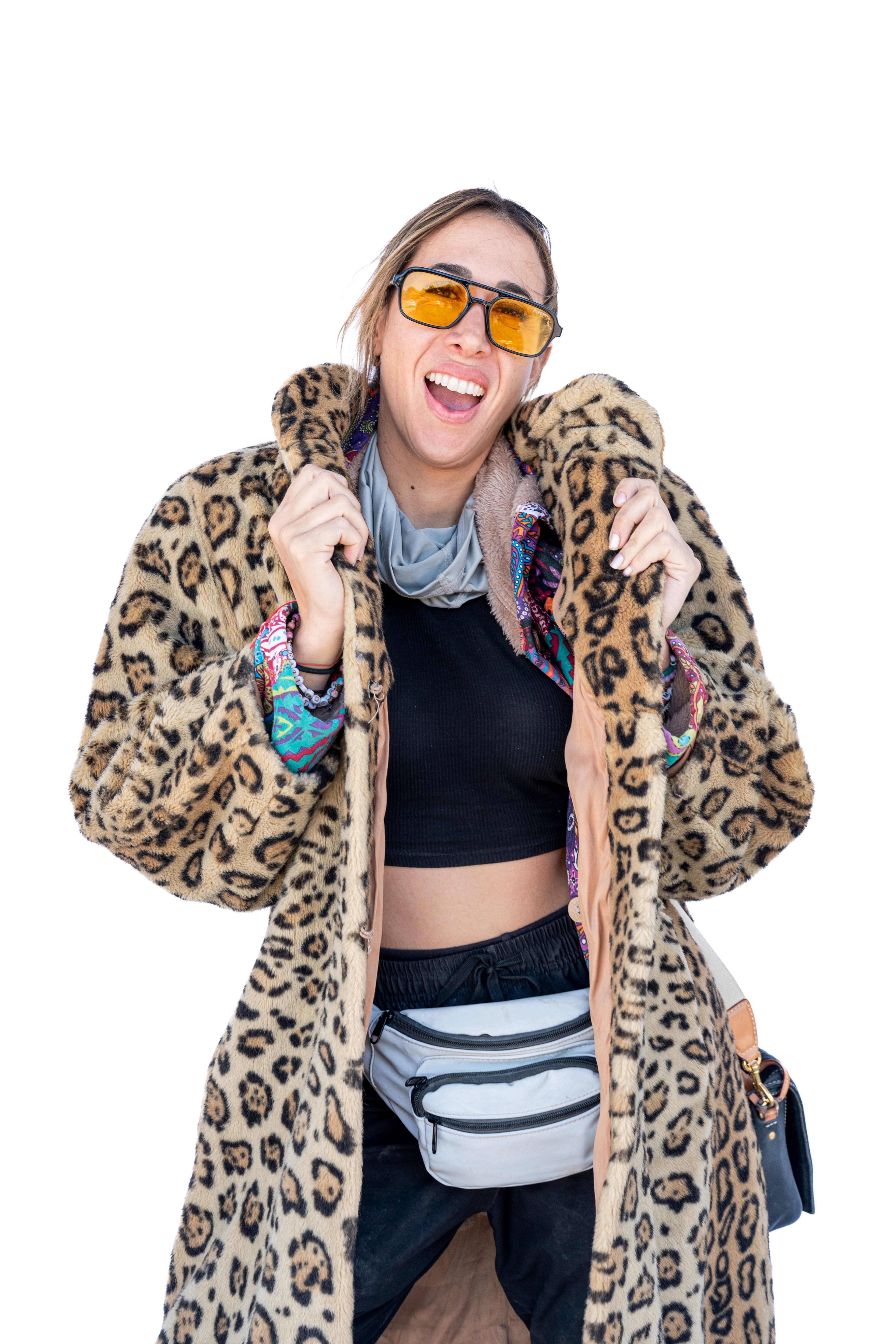 A person is smiling while wearing a leopard-print fur coat, yellow-tinted sunglasses, and a gray fanny pack. They also wear a black crop top and colorful accessories.