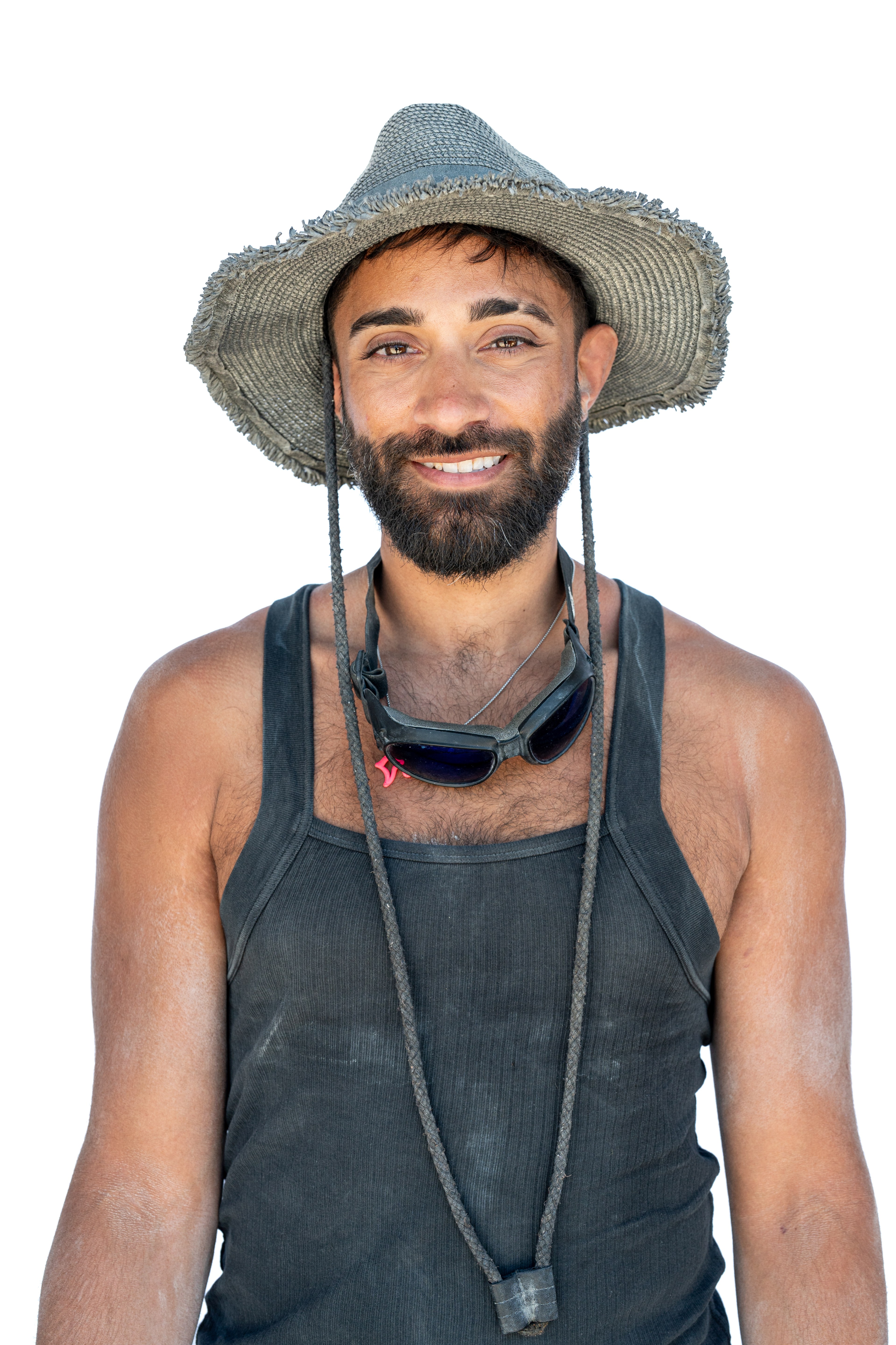 A smiling man with a beard is wearing a frayed hat, black tank top, and sunglasses around his neck. He is looking directly at the camera against a white background.