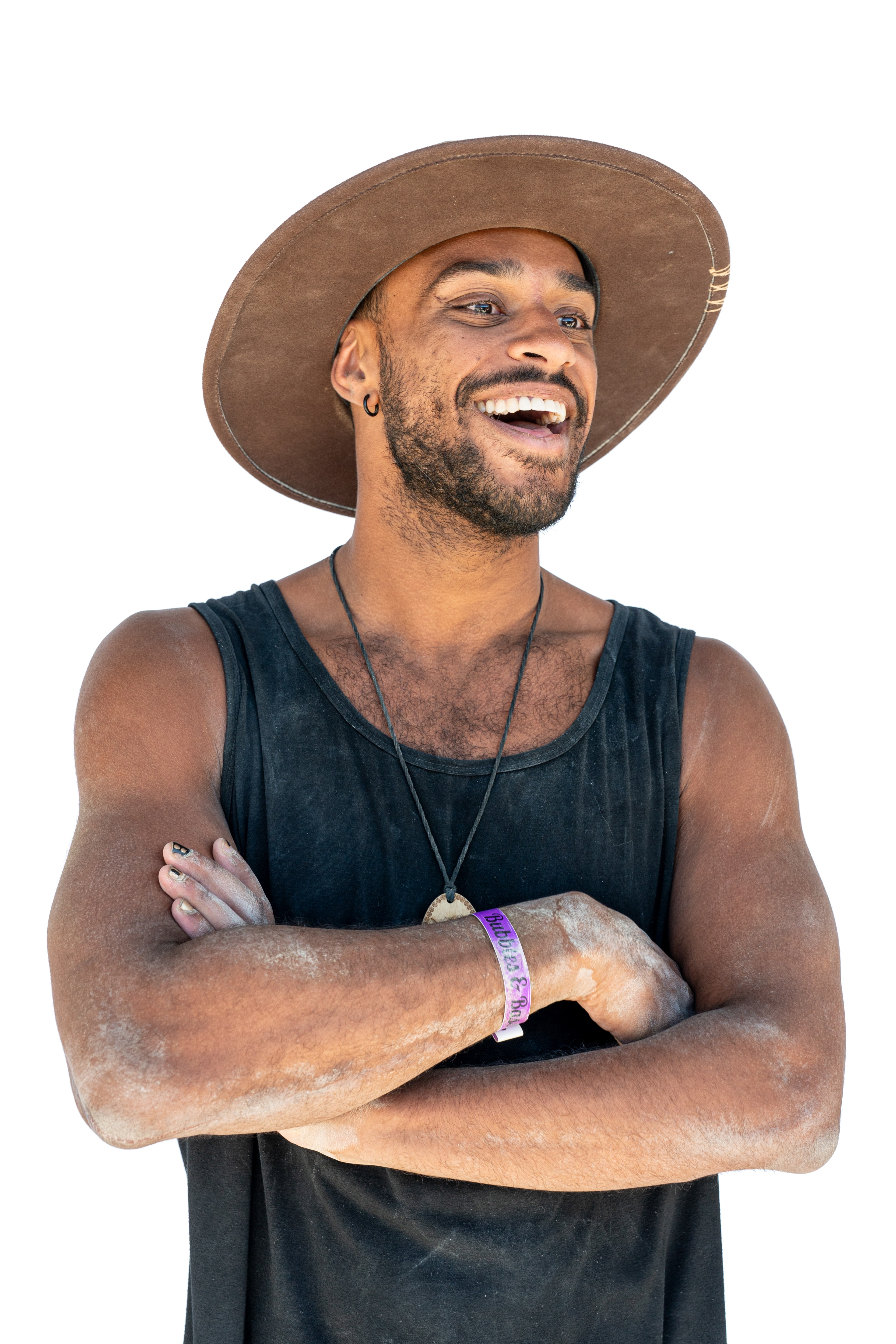 A person with a broad smile wears a wide-brimmed hat and a sleeveless black shirt. Their arms are crossed, and they have a bracelet and necklace.