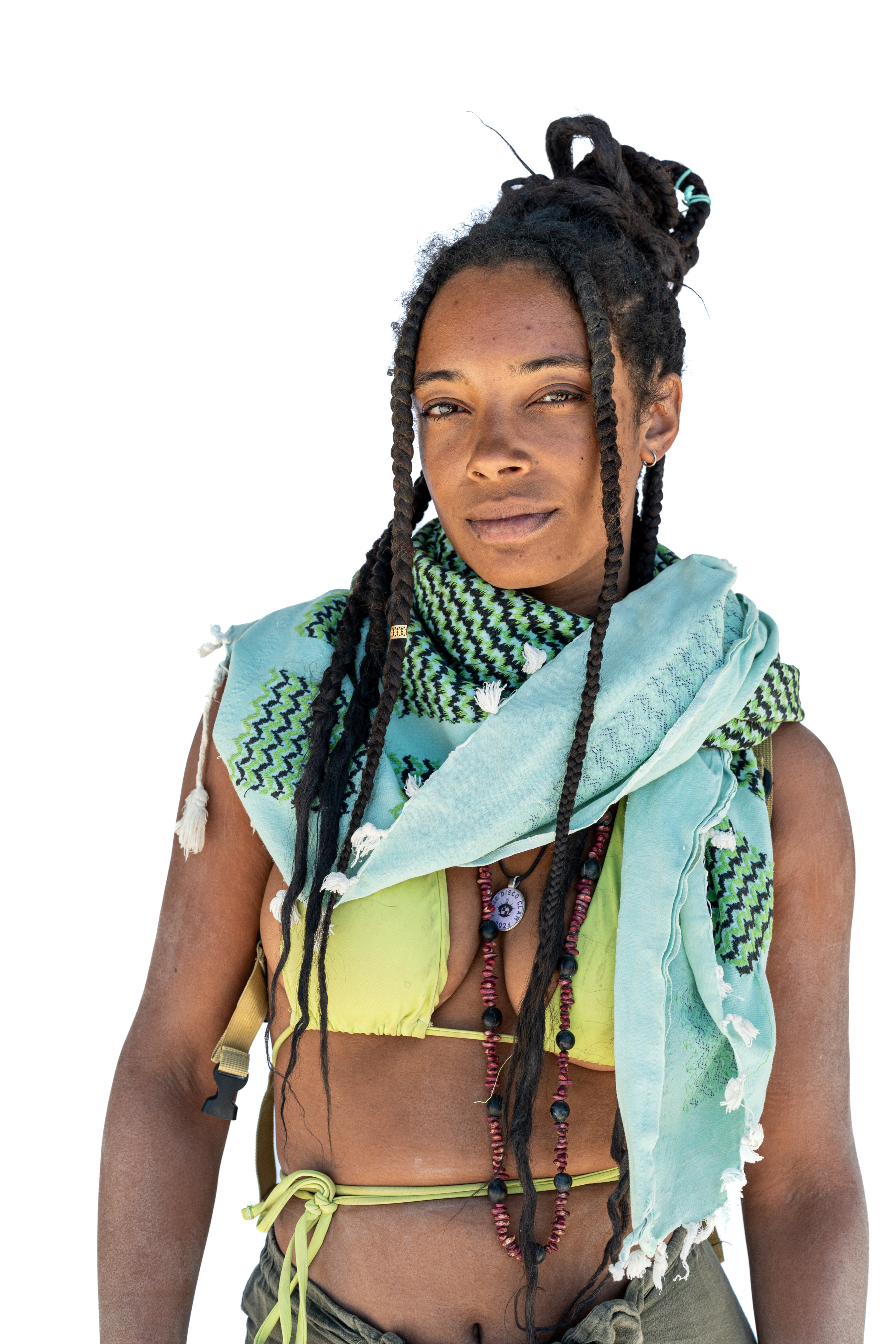 A person with long braids and a topknot hairstyle is wearing a green scarf with black patterns, a green bikini top, and a beaded necklace with a pendant.