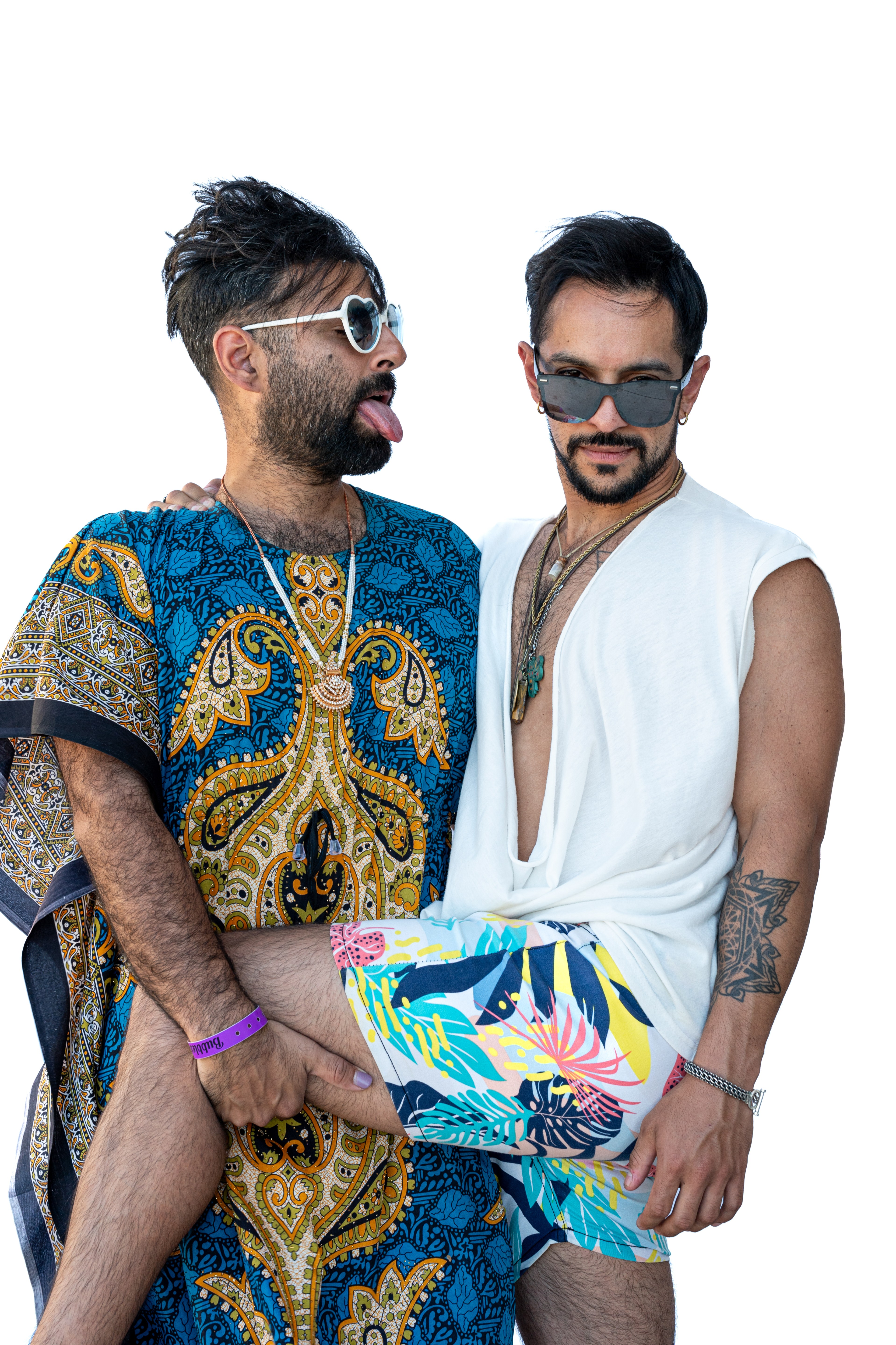 Two men stand close together, one in a colorful patterned robe and white sunglasses, playfully sticking out his tongue. The other wears a white sleeveless top and patterned shorts.