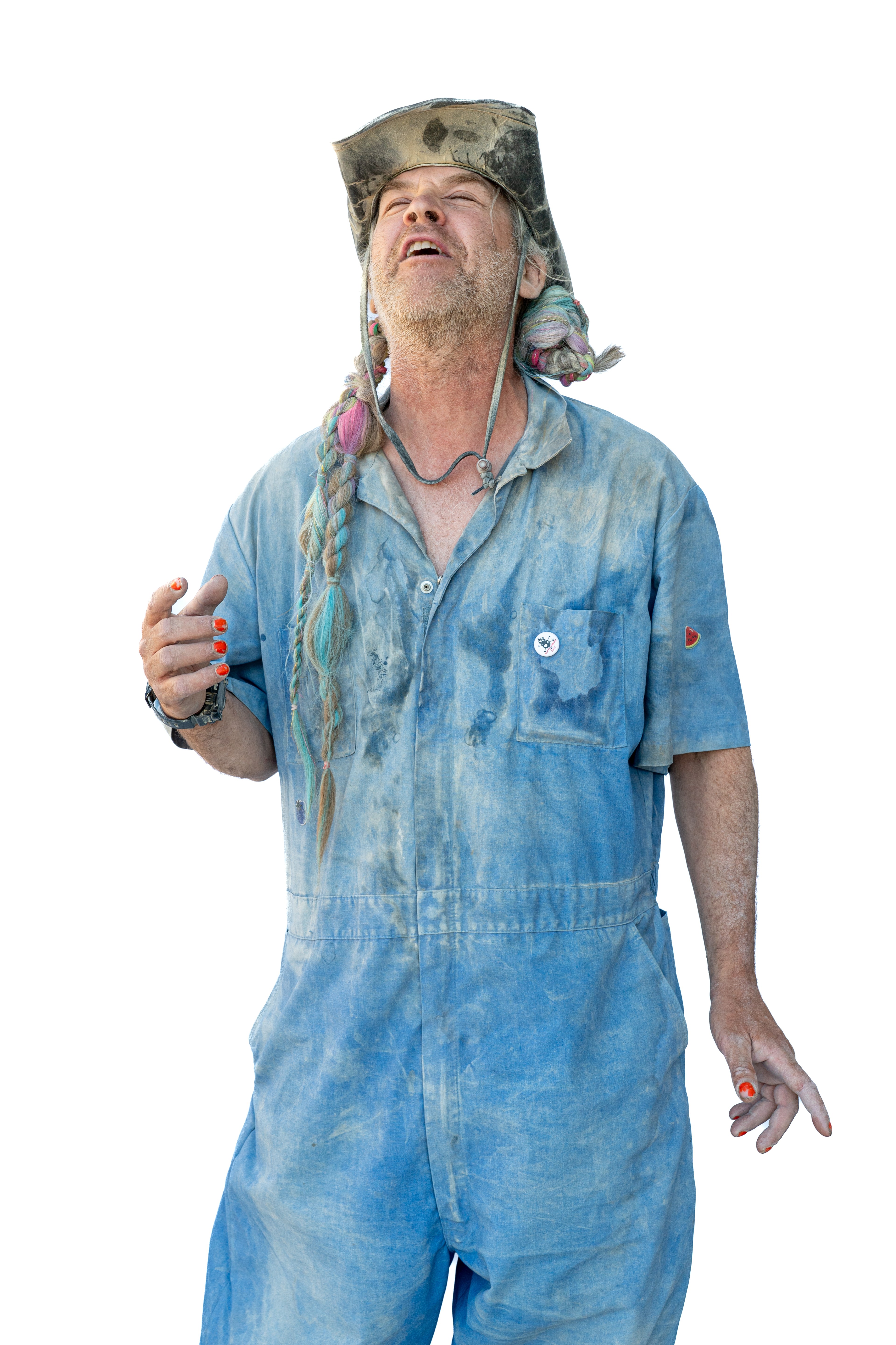 A bearded man in a dusty blue jumpsuit with braided, colorful hair is wearing a worn hat. He's holding his hand up, with an expression that seems thoughtful or wistful.