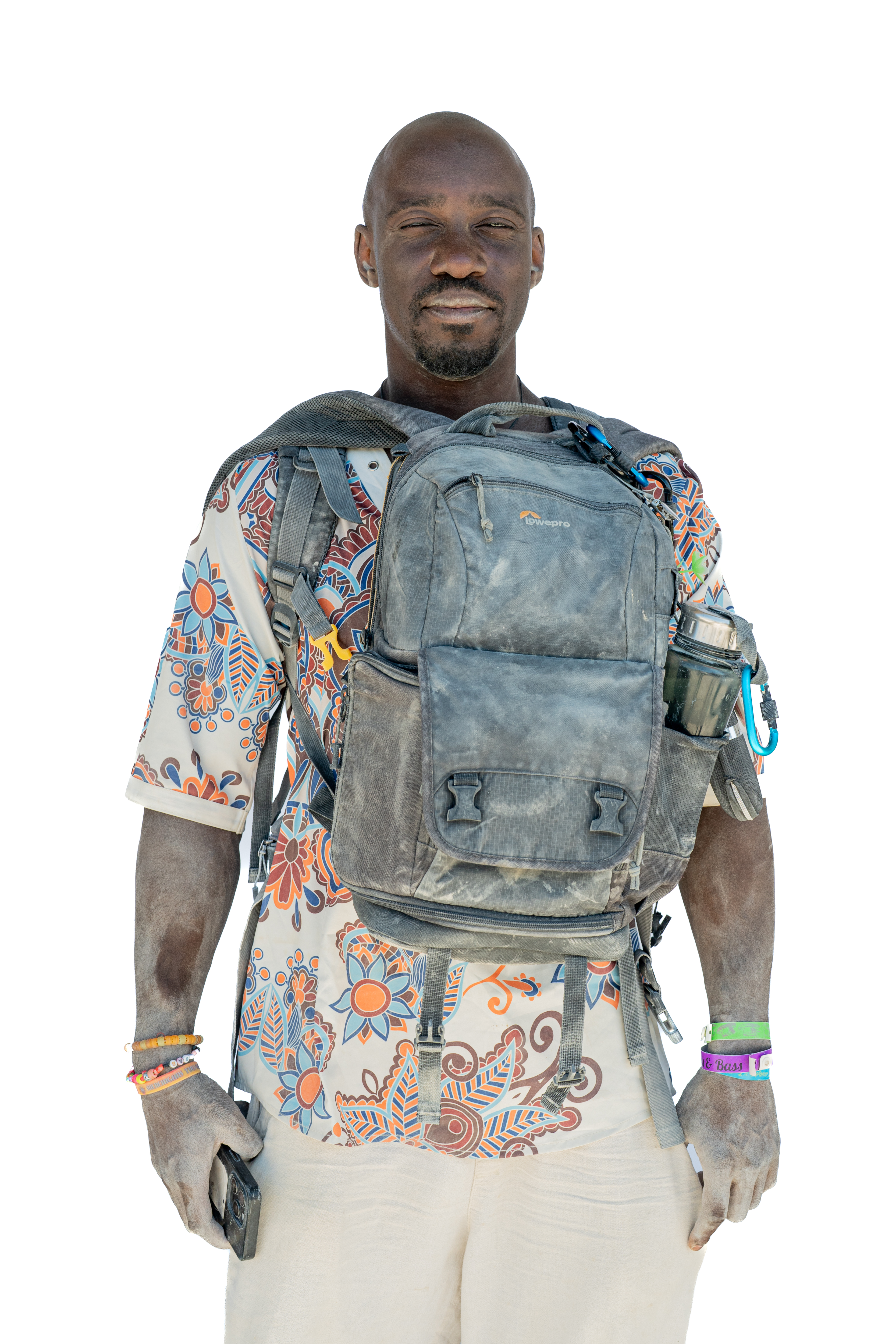 A person wearing a colorful, patterned shirt and white pants carries a dusty backpack with a water bottle. They have various bracelets on their wrist and hold a phone.