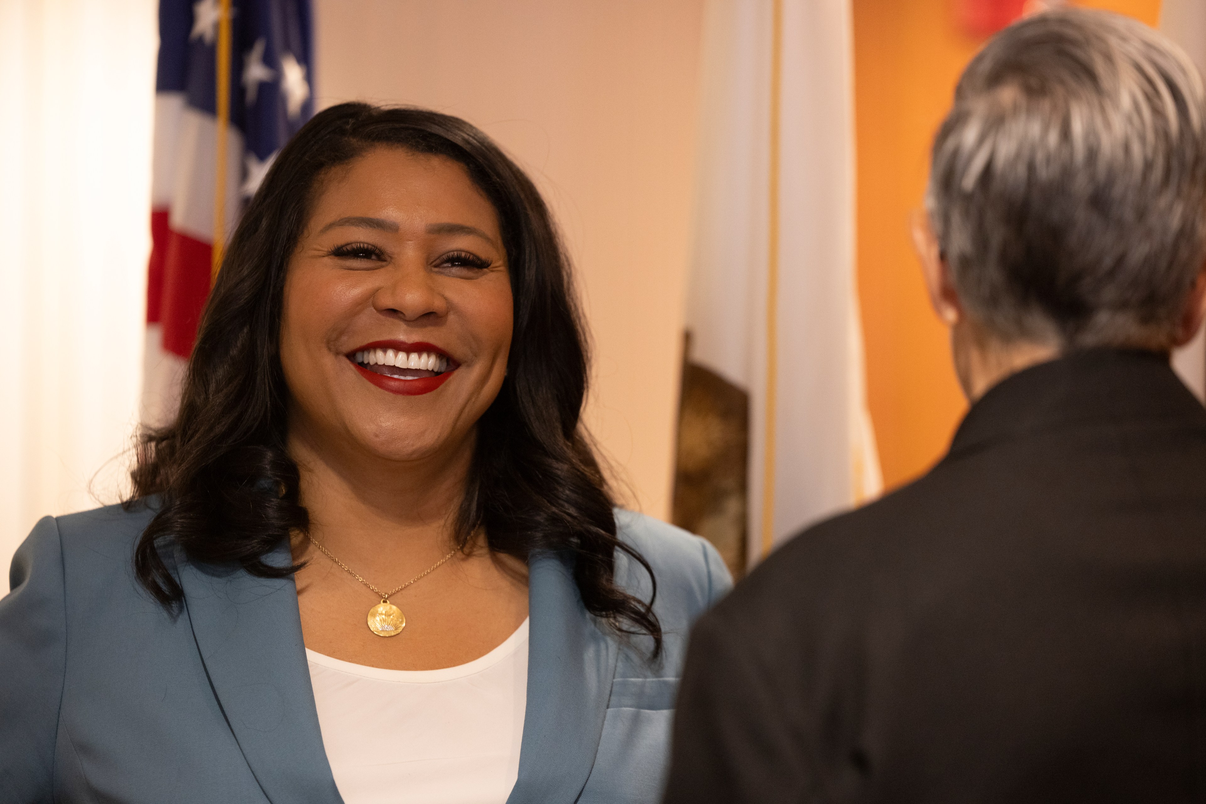 A woman in a blue blazer and white top smiles brightly at someone. Flags are visible in the background, suggesting the setting might be a formal or civic event.