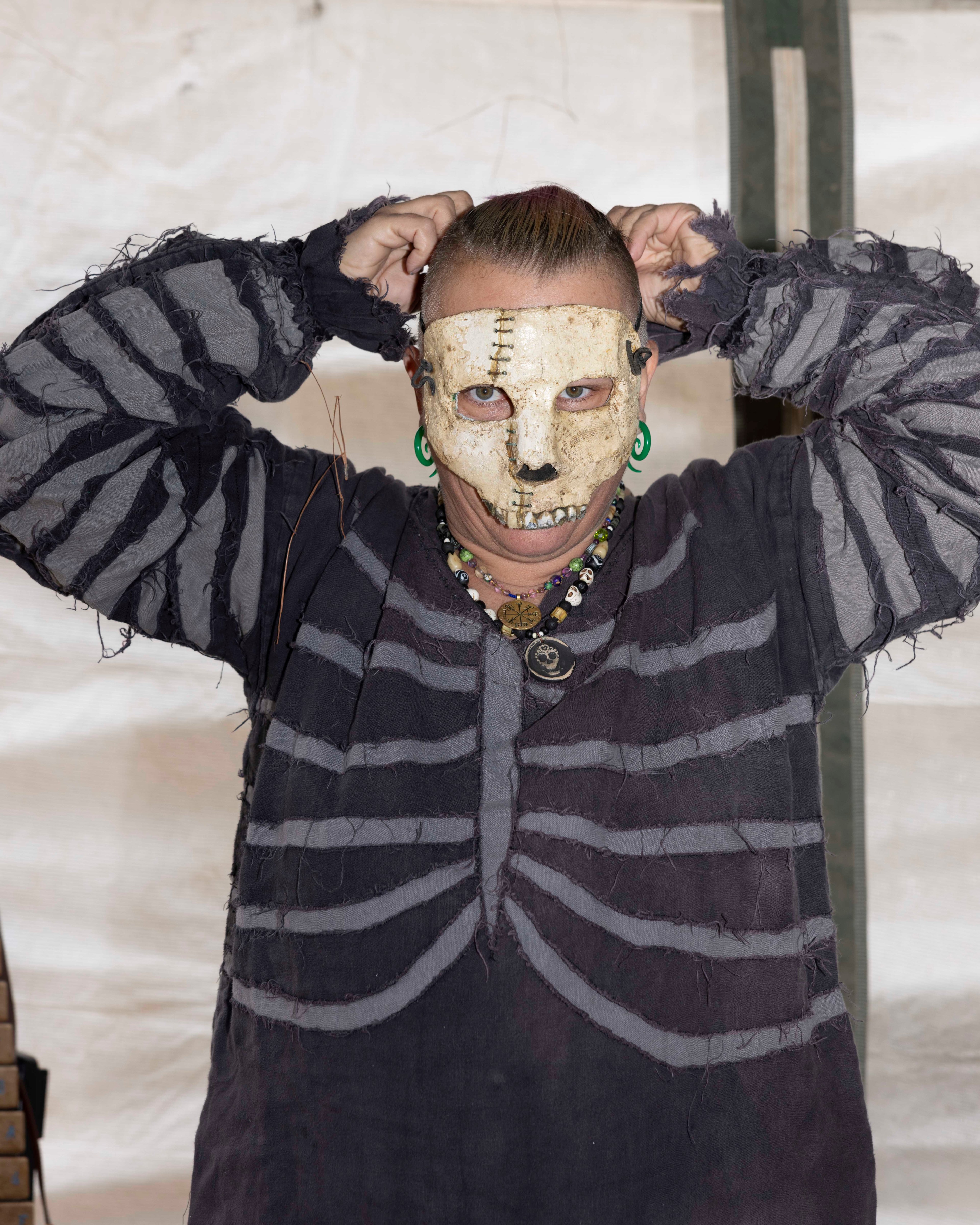 The individual is wearing a skeleton-themed costume with a stitched white skull mask and adjusting their hair. They also have green ear plugs and multiple necklaces.