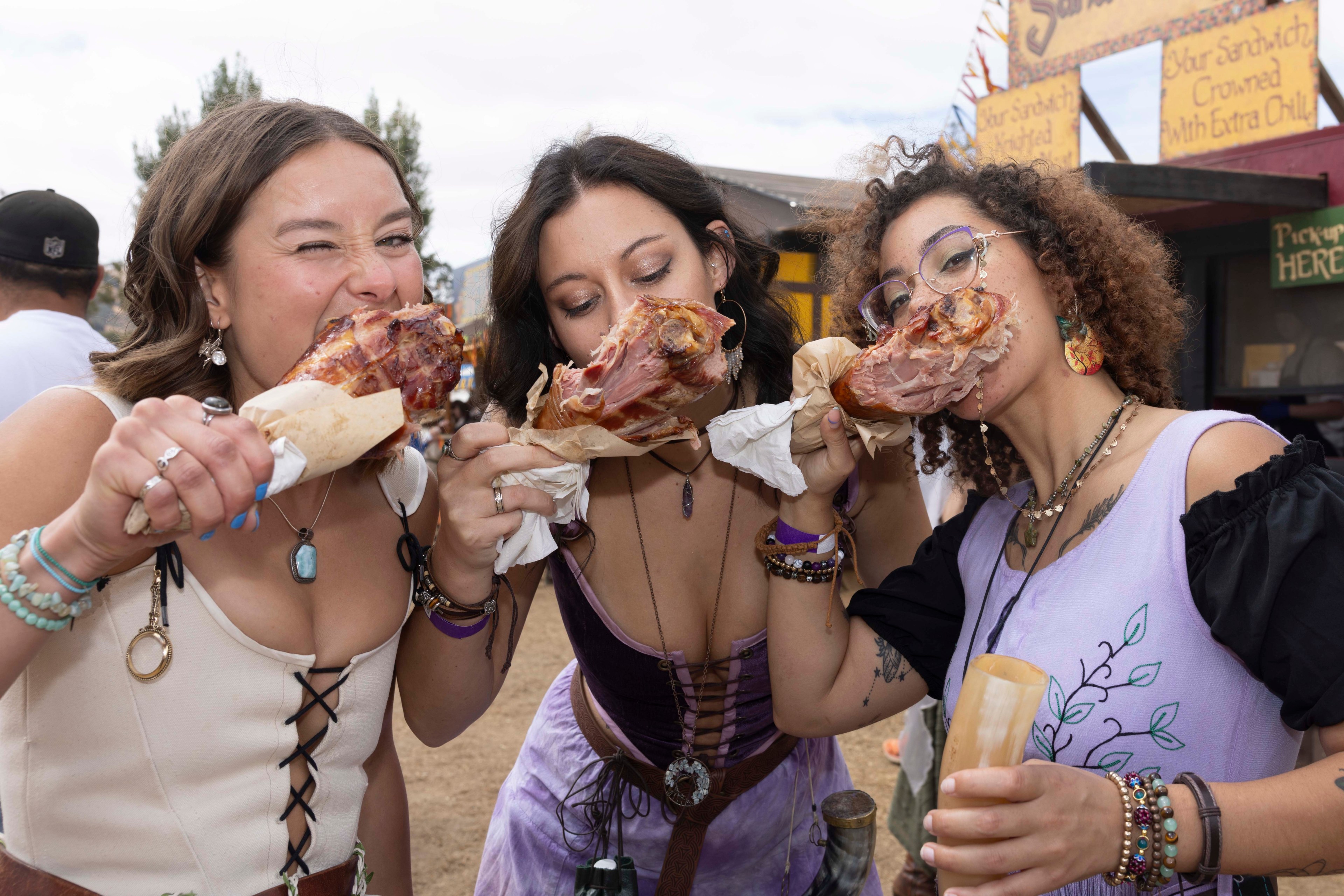 Three women dressed in colorful, Renaissance-style outfits are joyfully biting into large turkey legs at an outdoor festival.