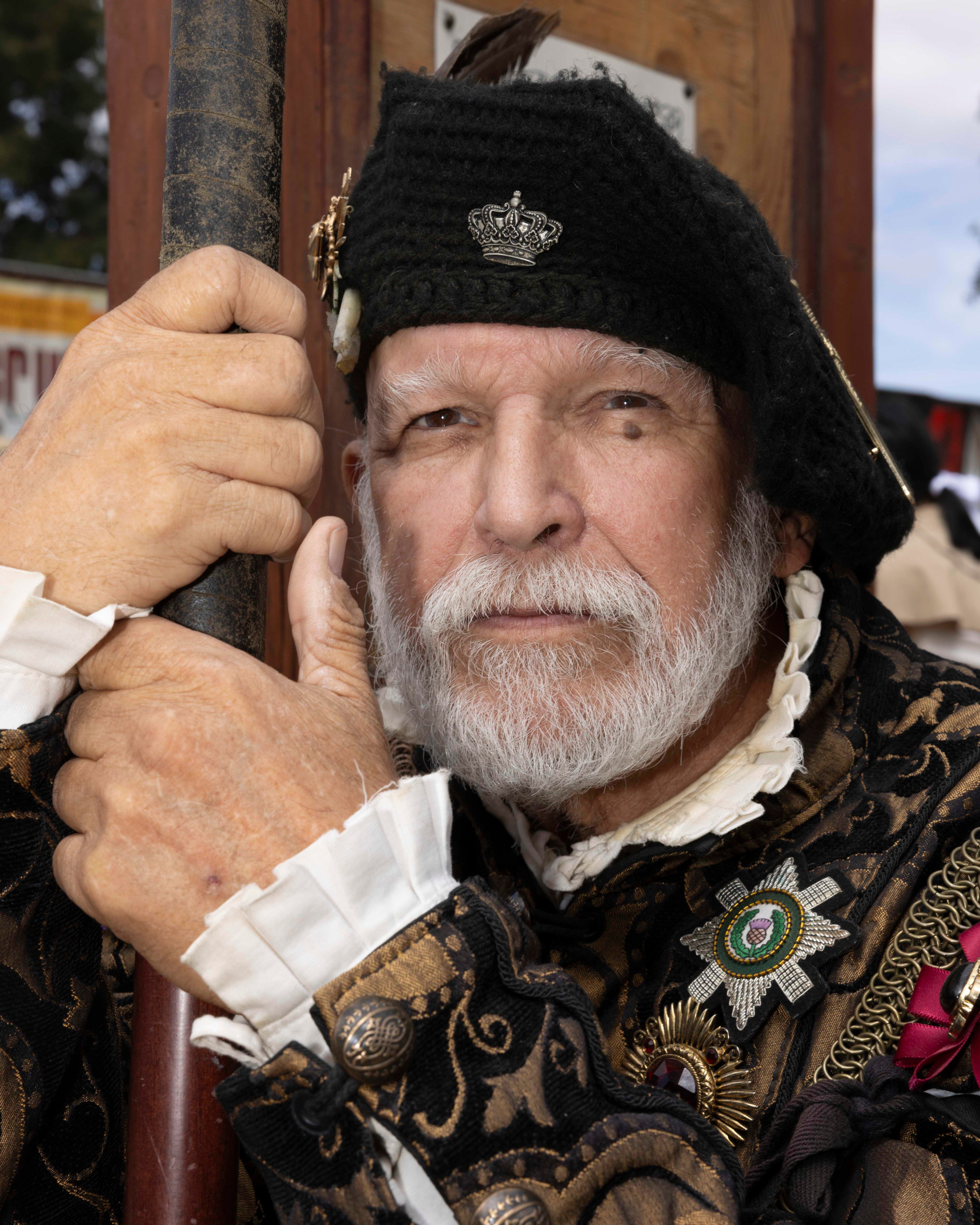 A bearded man in ornate armor and a decorative hat gazes intently while holding a staff, featuring a detailed pattern and medals on his costume.