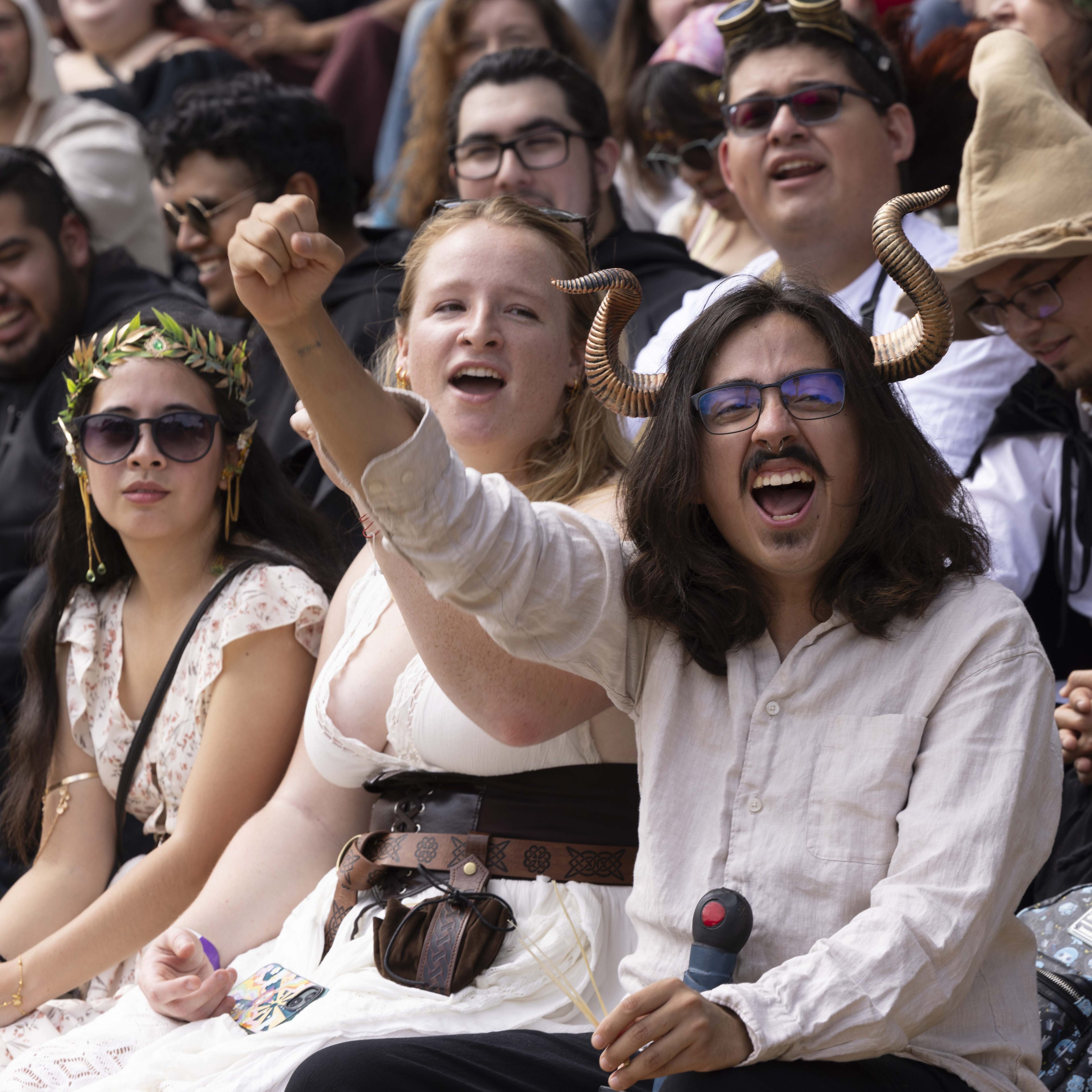 A cheering crowd in costumes, including a person with horn headgear raising a fist enthusiastically, others wear crowns, glasses, and various themed outfits.