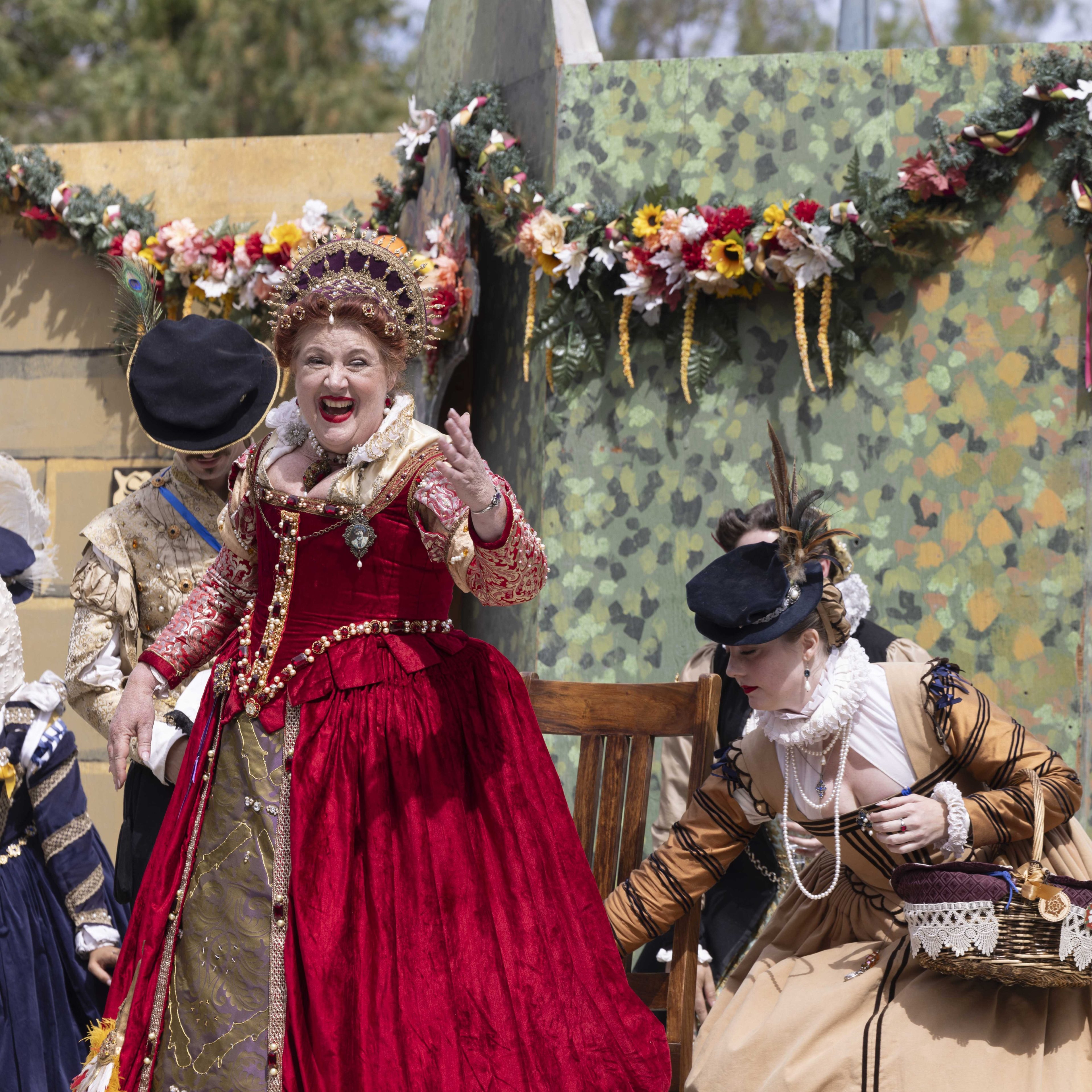 A woman in a lavish red gown and crown speaks animatedly, while another in a historical costume bends over near a wooden chair, against a decorated wall backdrop.