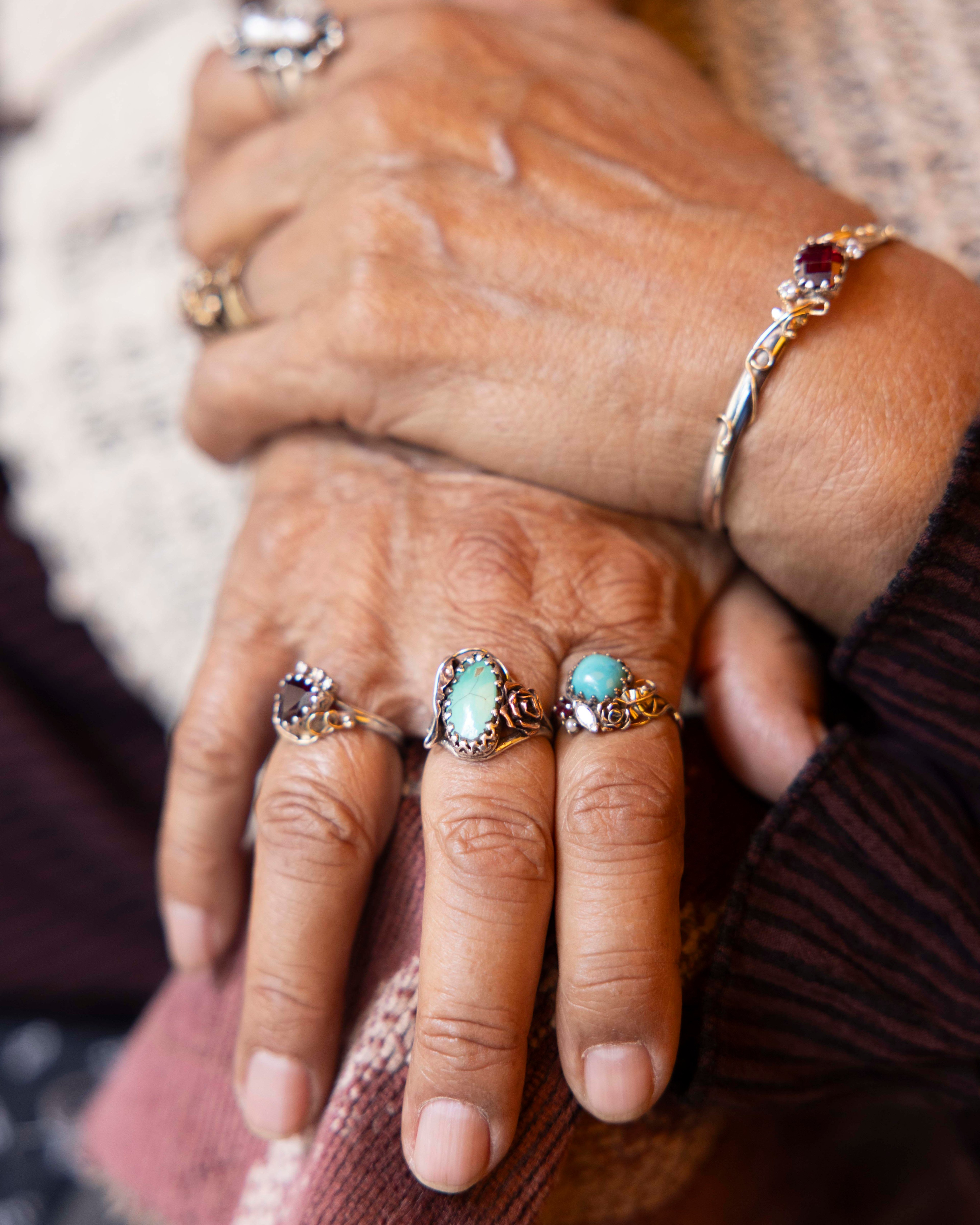 The image shows a close-up of a person's hands folded together, adorned with several rings featuring turquoise stones and intricate designs, and a bracelet with a dark red gem.