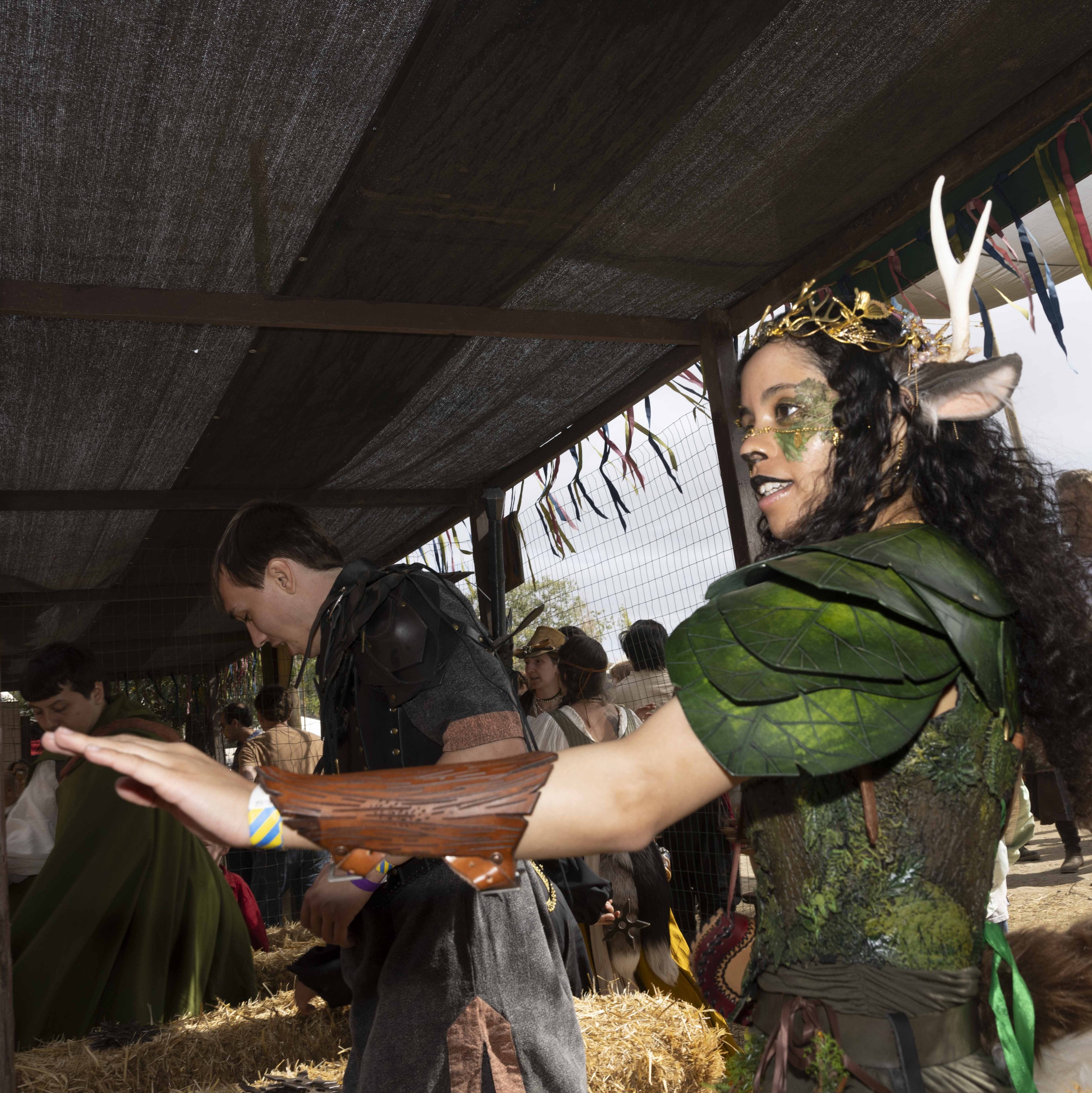 A woman in a vibrant, leafy costume with antlers and face paint participates in a rustic, possibly fantasy-themed event with hay and other similarly dressed individuals.