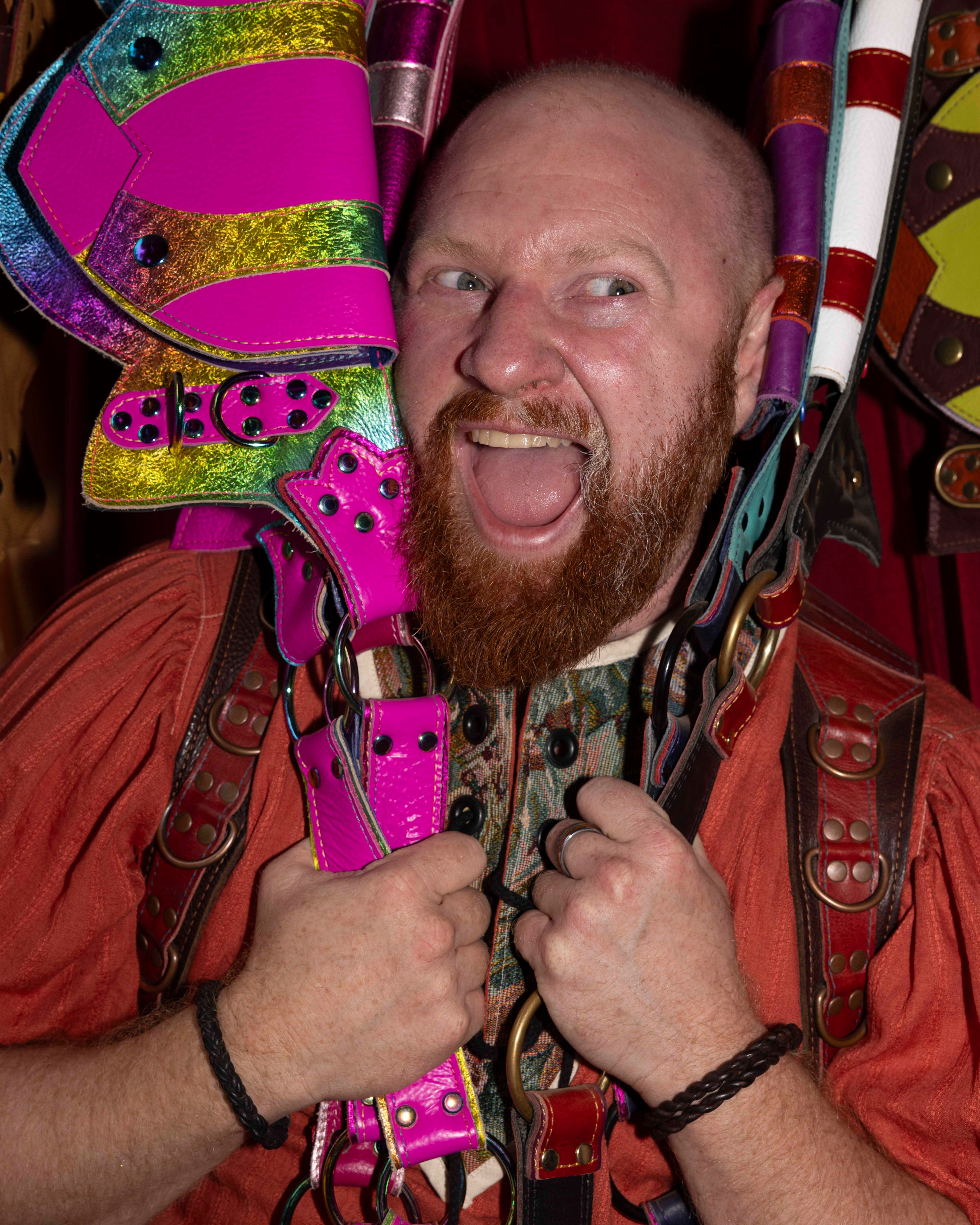 A man with a red beard grins widely while holding colorful, shiny leather straps, including pink and rainbow ones, against his head and shoulders.