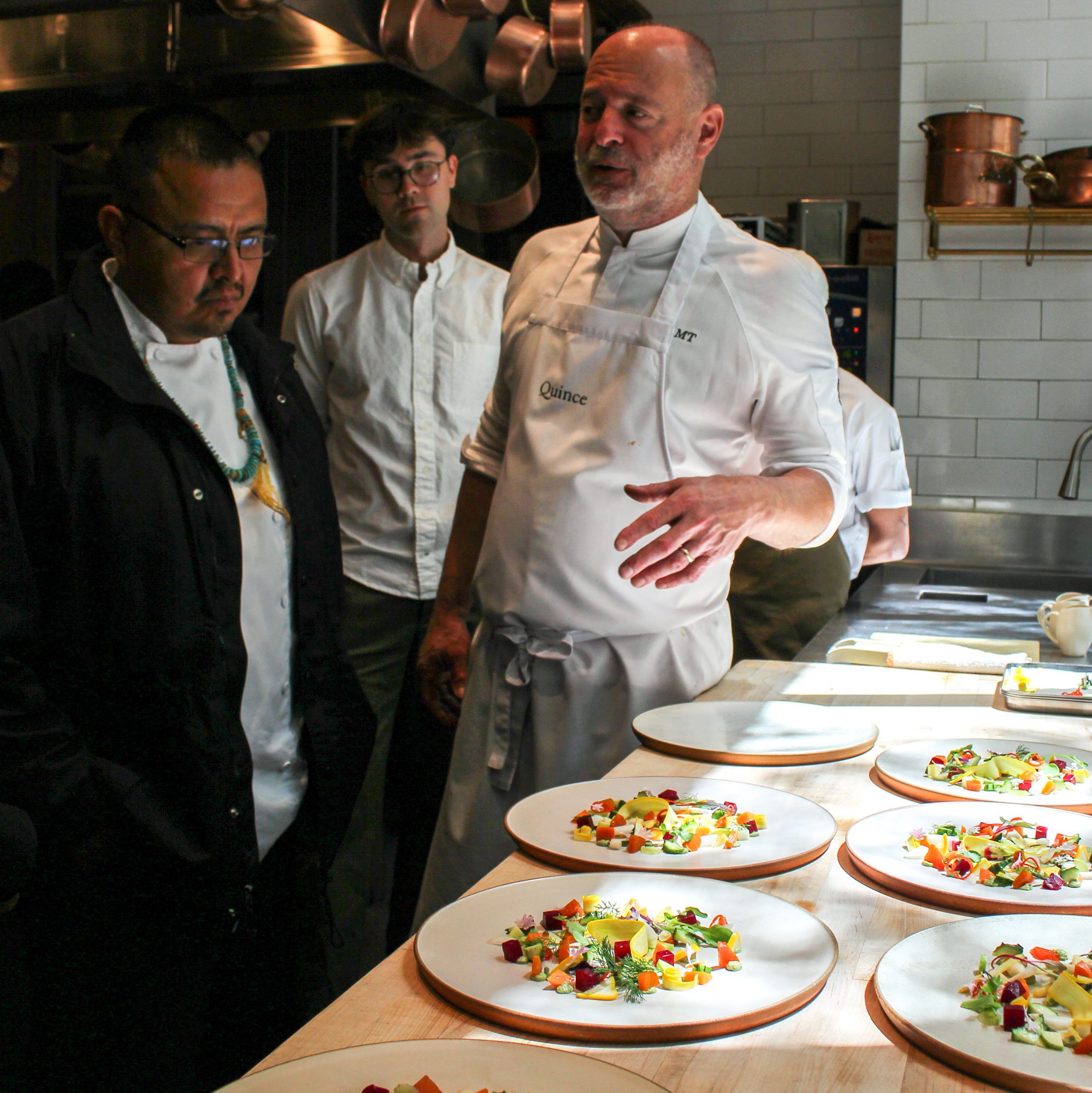 A chef in a white apron is explaining something to others in a kitchen, with several plates of colorful, finely arranged food on a wooden table in front of them.