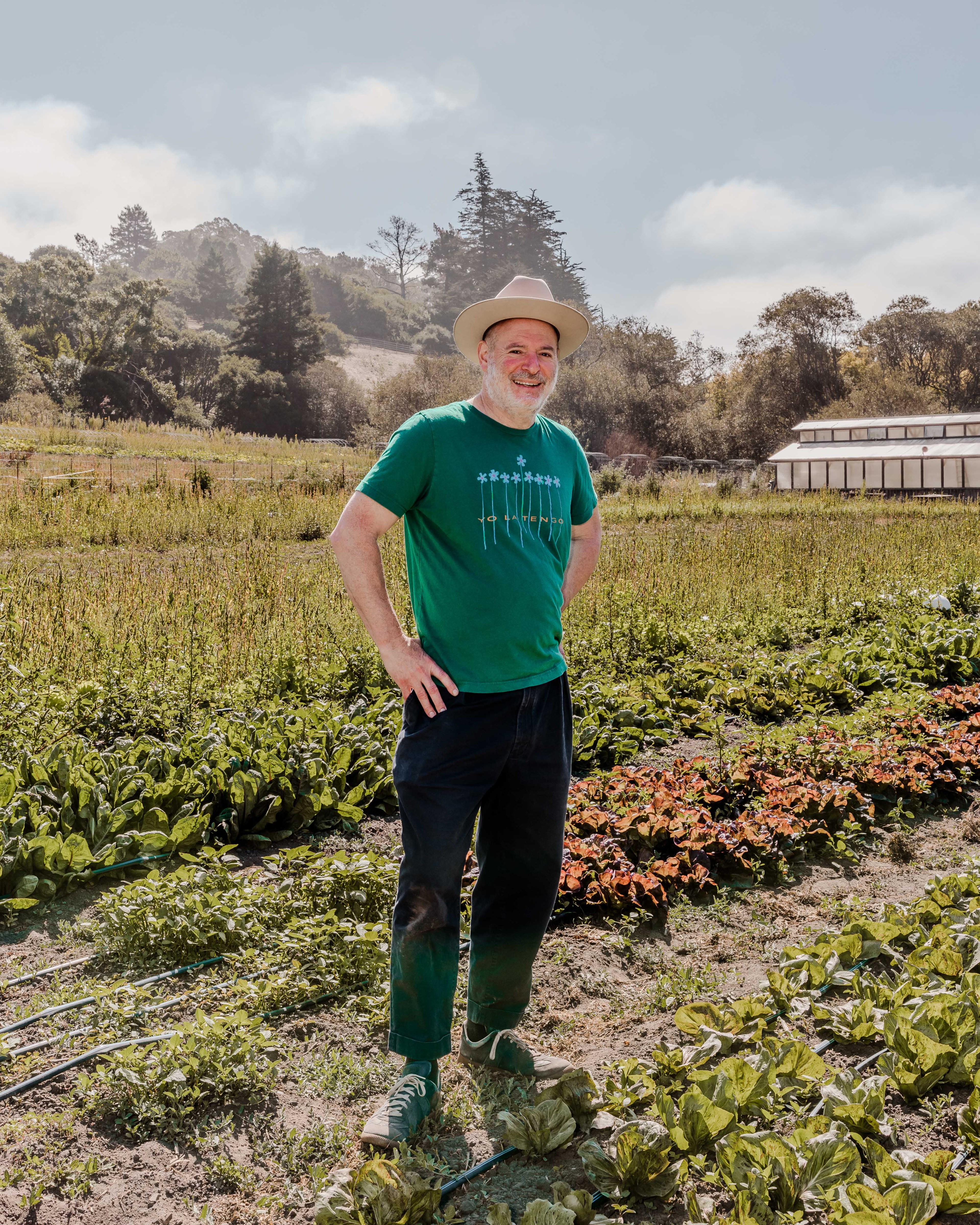 A smiling man stands in a lush vegetable garden, wearing a green T-shirt, dark pants, green shoes, and a beige hat. Trees and a building are visible in the background.