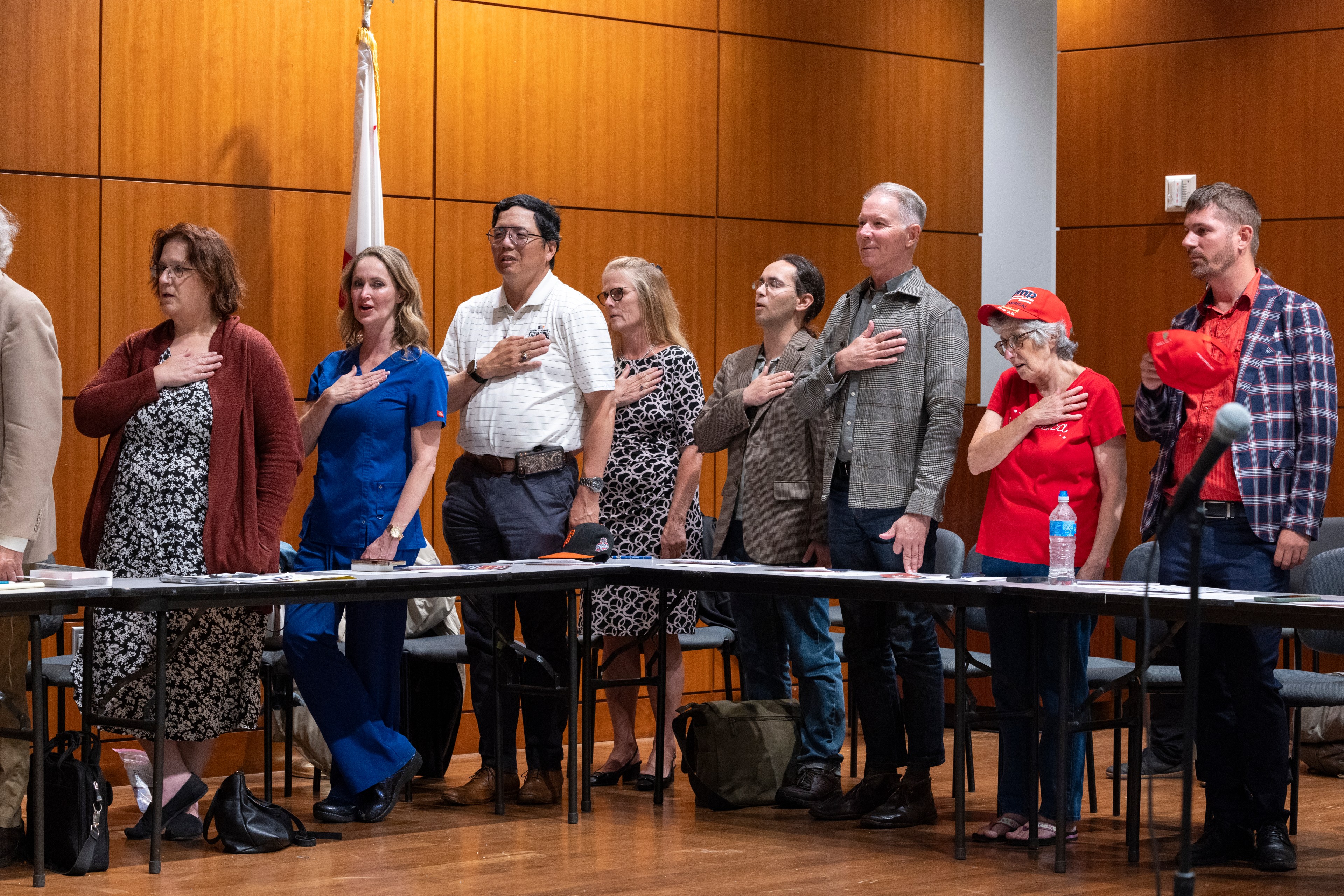 A group of nine people stands in a row with their right hands over their hearts in a wood-paneled room, possibly reciting the Pledge of Allegiance.