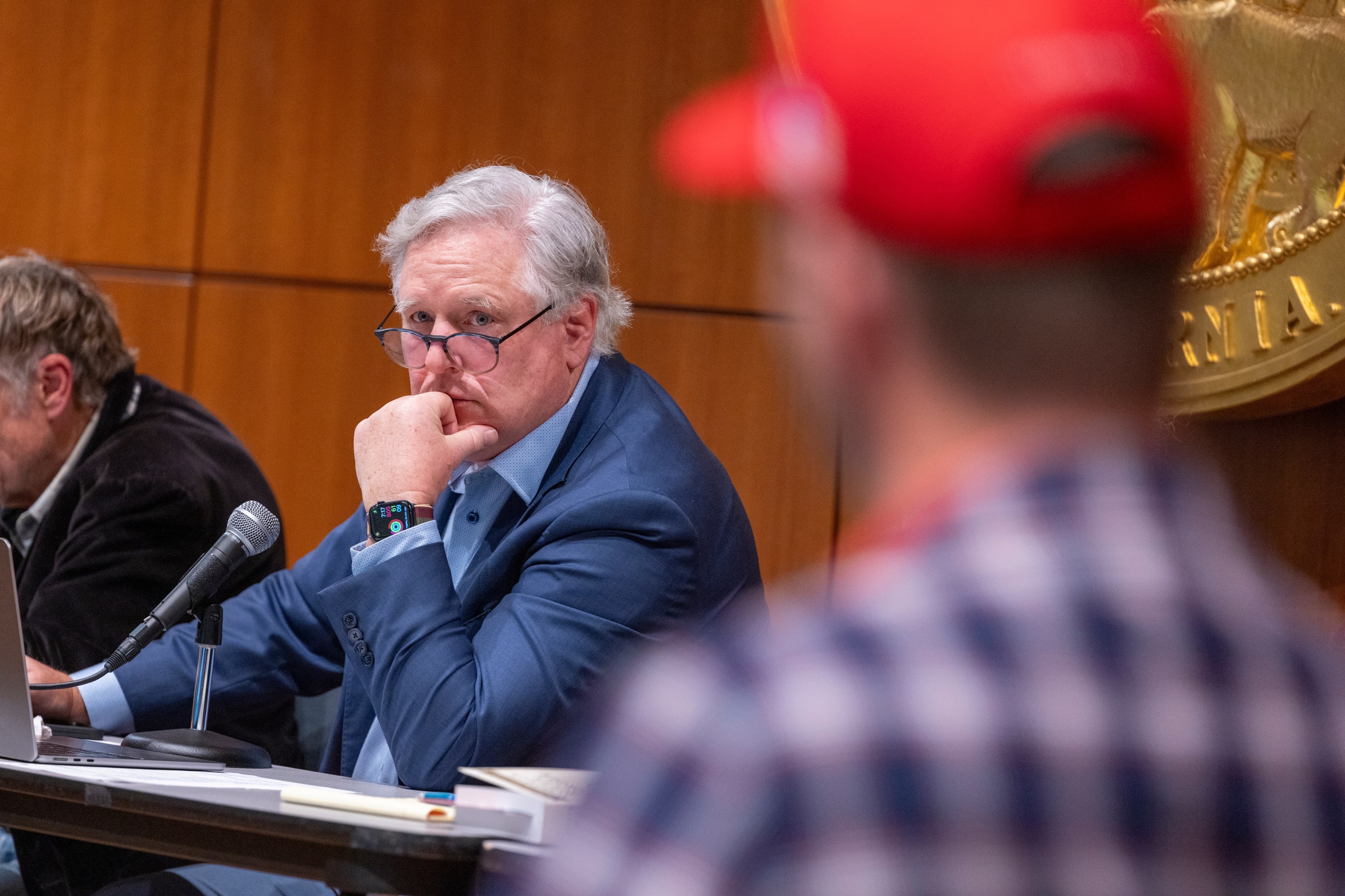 A man in a blue suit and glasses sits at a table, focusing intensely, with his hand on his chin. Another, blurred man in a red cap, is in the foreground.