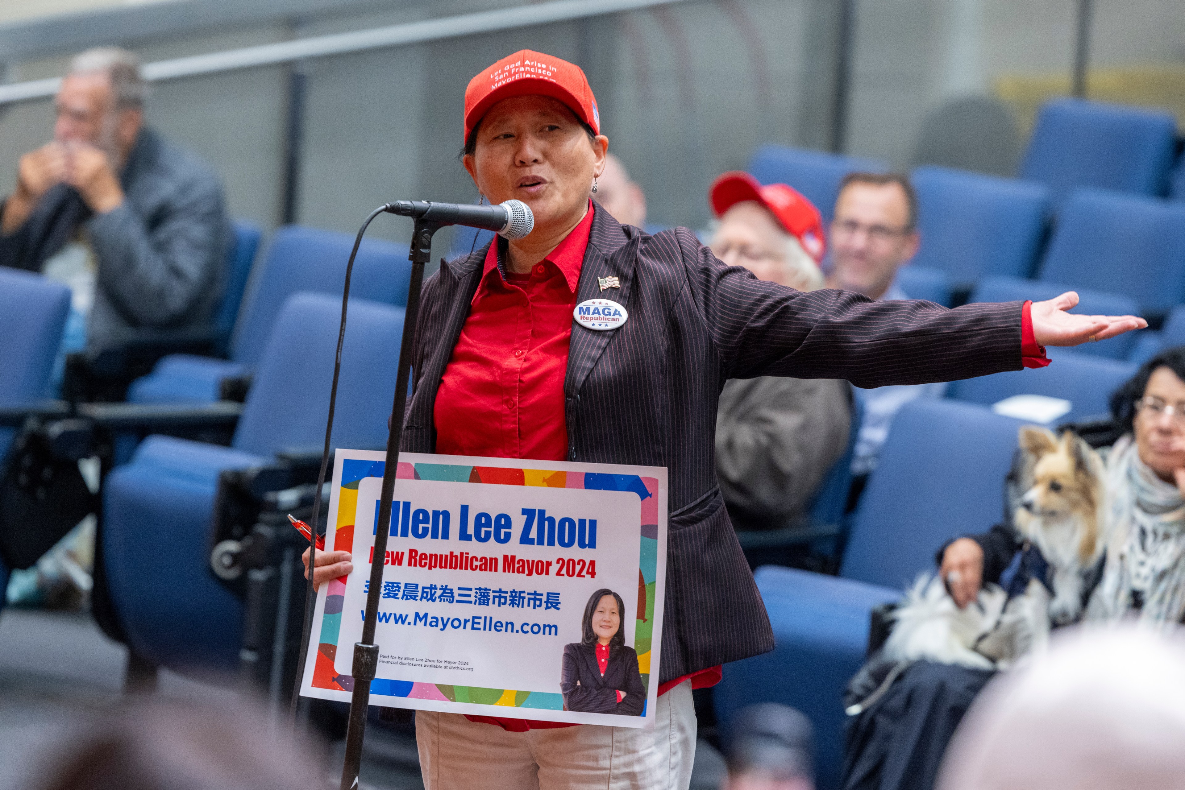 A woman in a red cap and shirt speaks into a microphone, holding a campaign sign for Ellen Lee Zhou, in a room with blue seats and an audience.