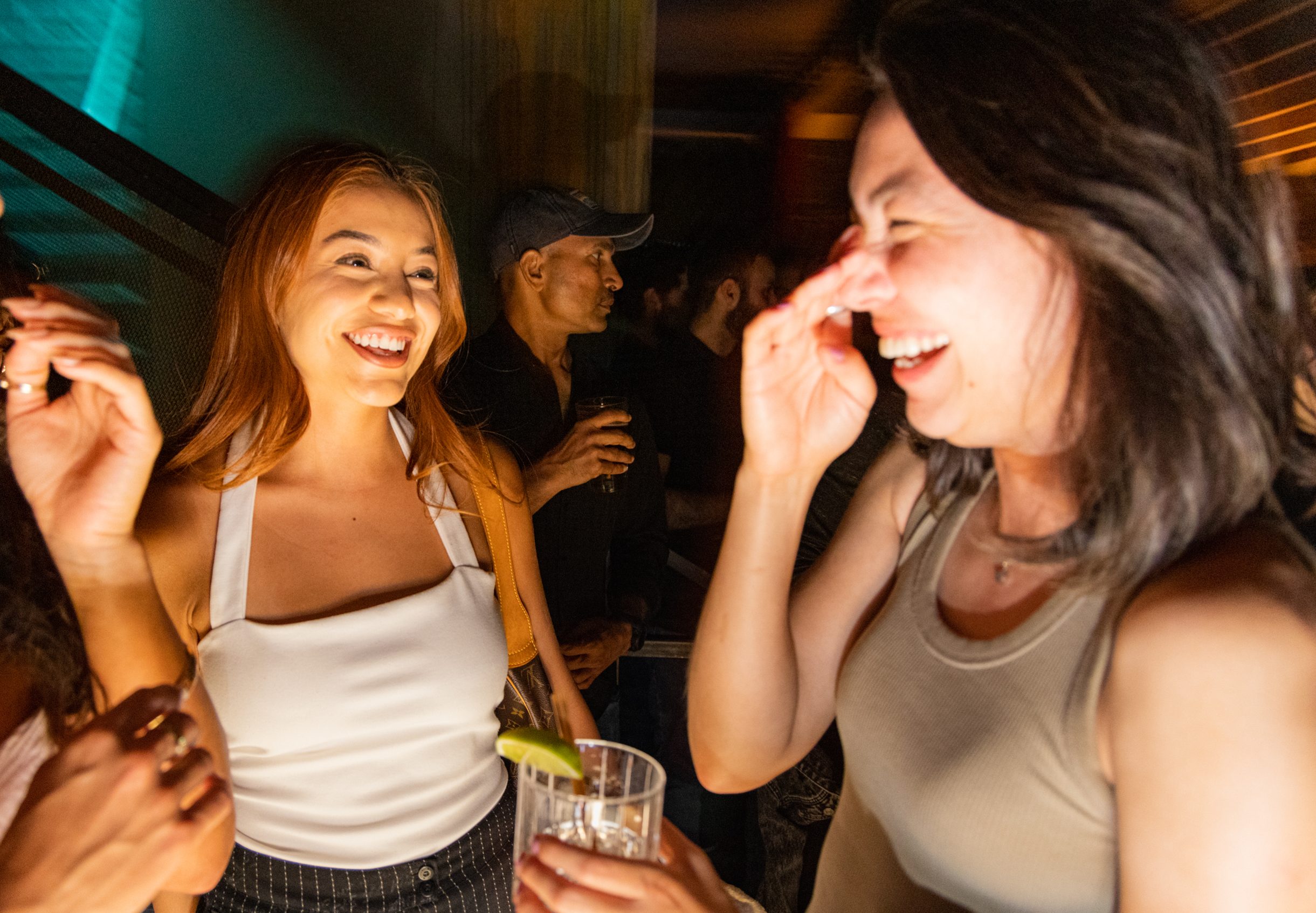 Three women are laughing and chatting in a dimly lit bar, with one holding a drink. A man in a cap stands behind them, holding a drink and observing the scene.