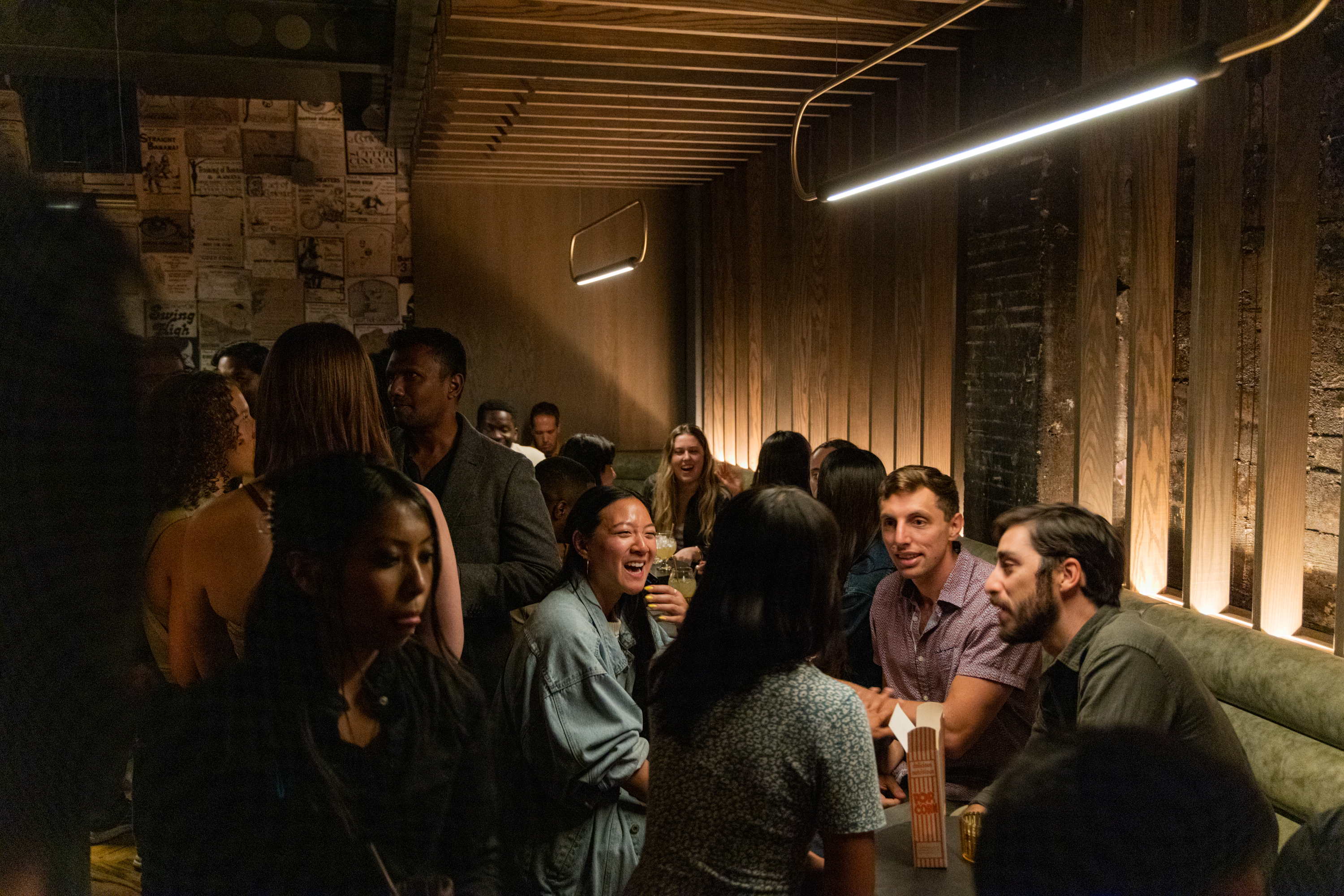 A dimly lit bar is filled with people engaged in conversations and laughter. Some sit around tables while others stand, creating a lively and warm atmosphere.