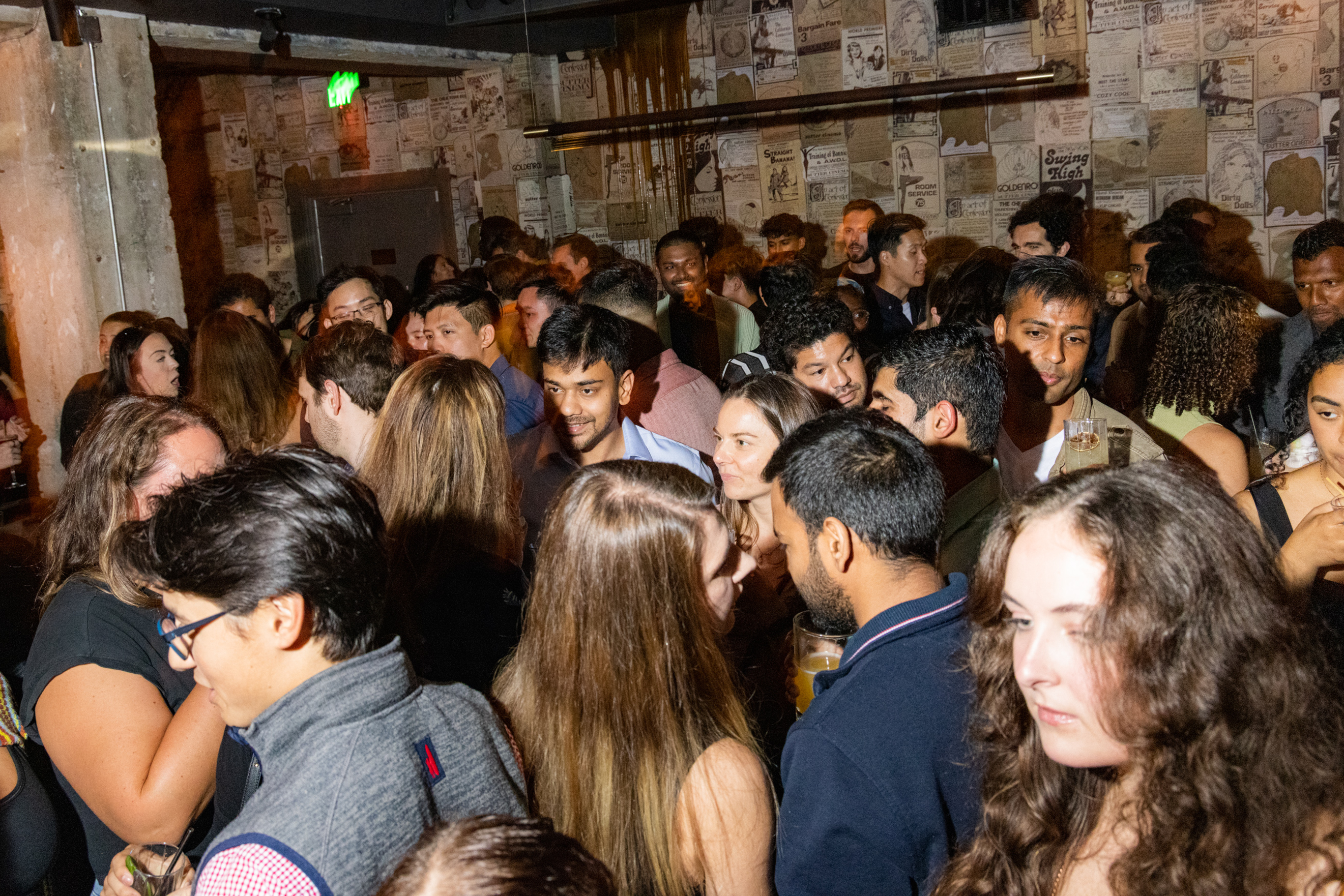 A crowded indoor gathering shows people mingling and conversing. The walls are covered in vintage posters, and some attendees are holding drinks, creating a lively atmosphere.