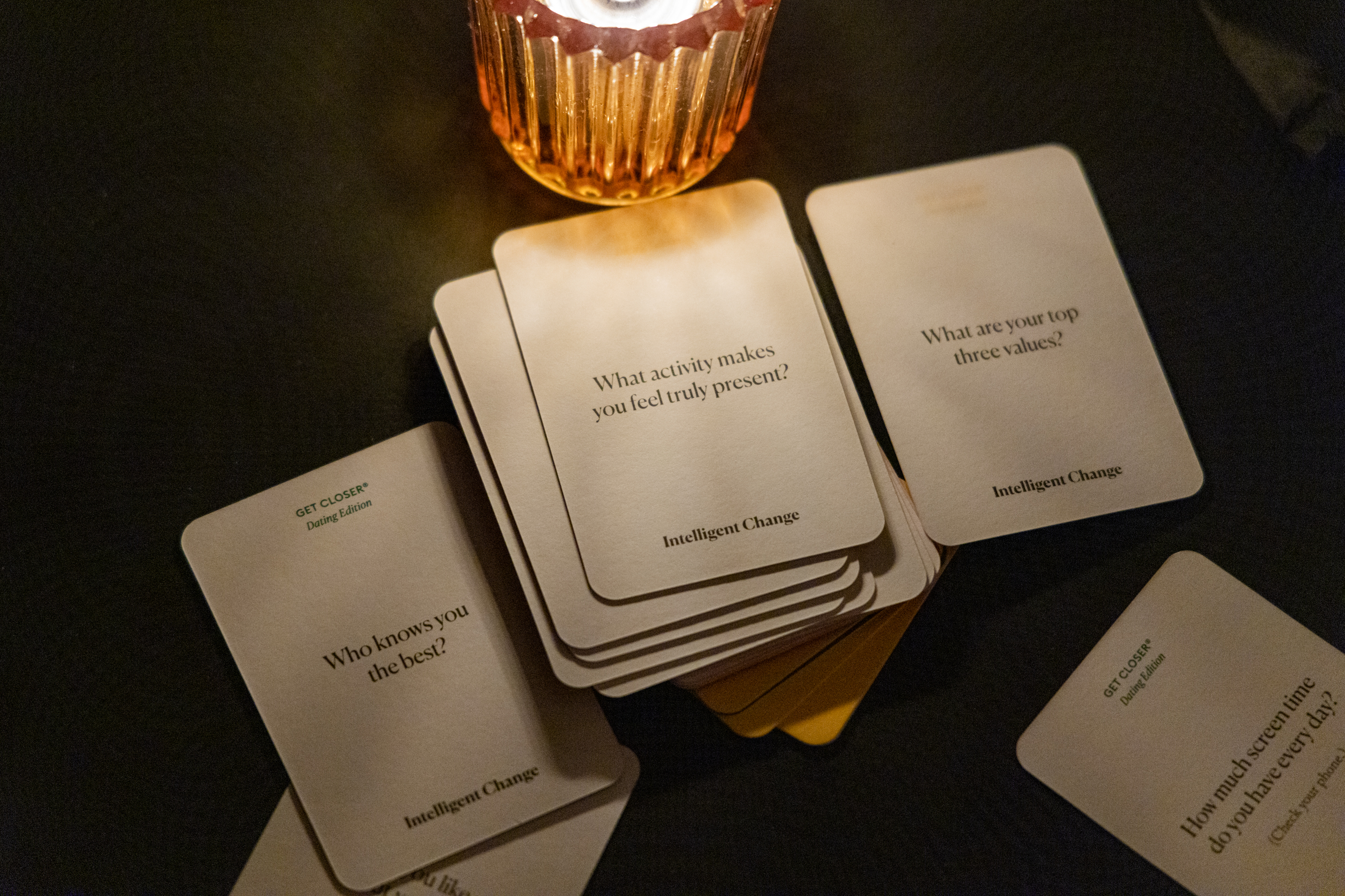 A stack of “Get Closer” dating edition cards with questions like “What activity makes you feel truly present?” is illuminated by a small candle in a glass holder.