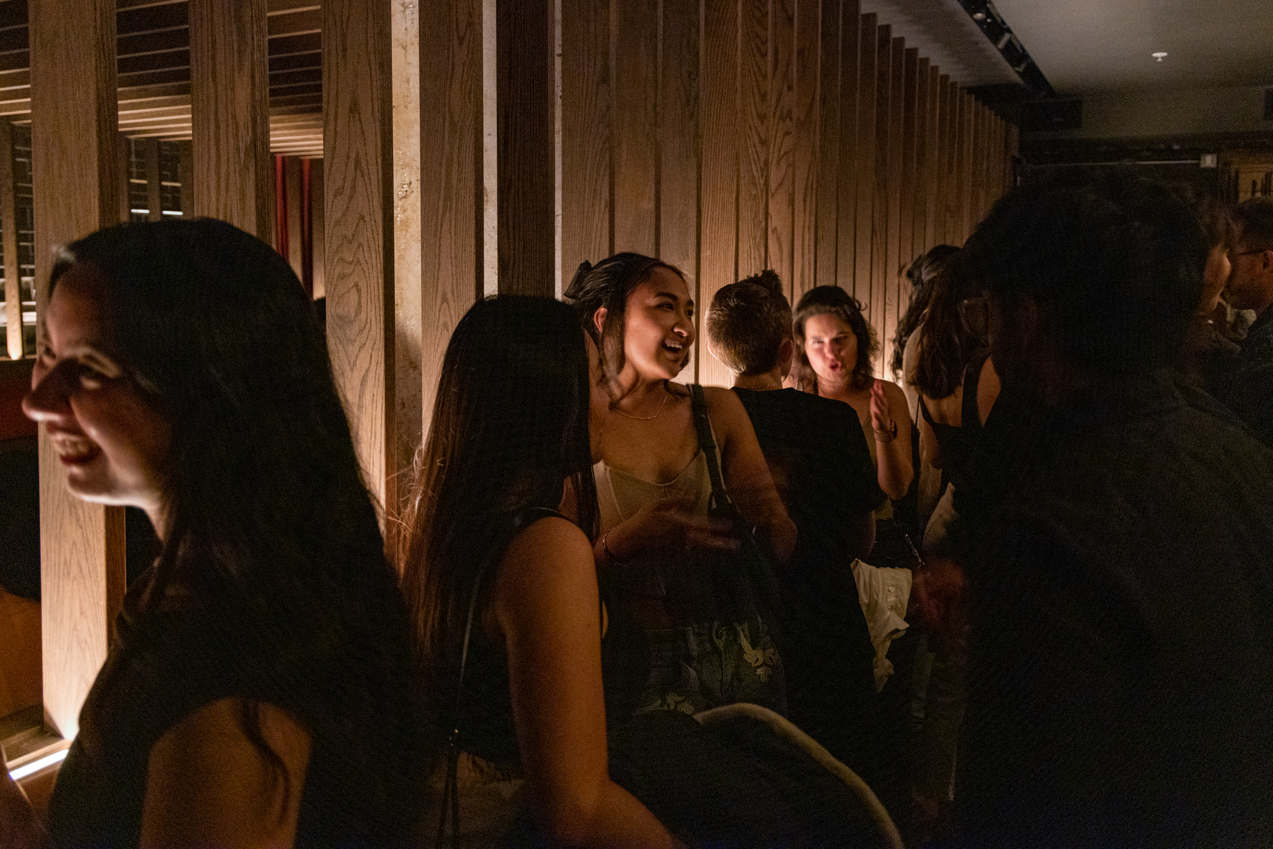 A group of people socialize in a dimly lit room with wooden panel walls, engaged in animated conversations and appearing to enjoy themselves.