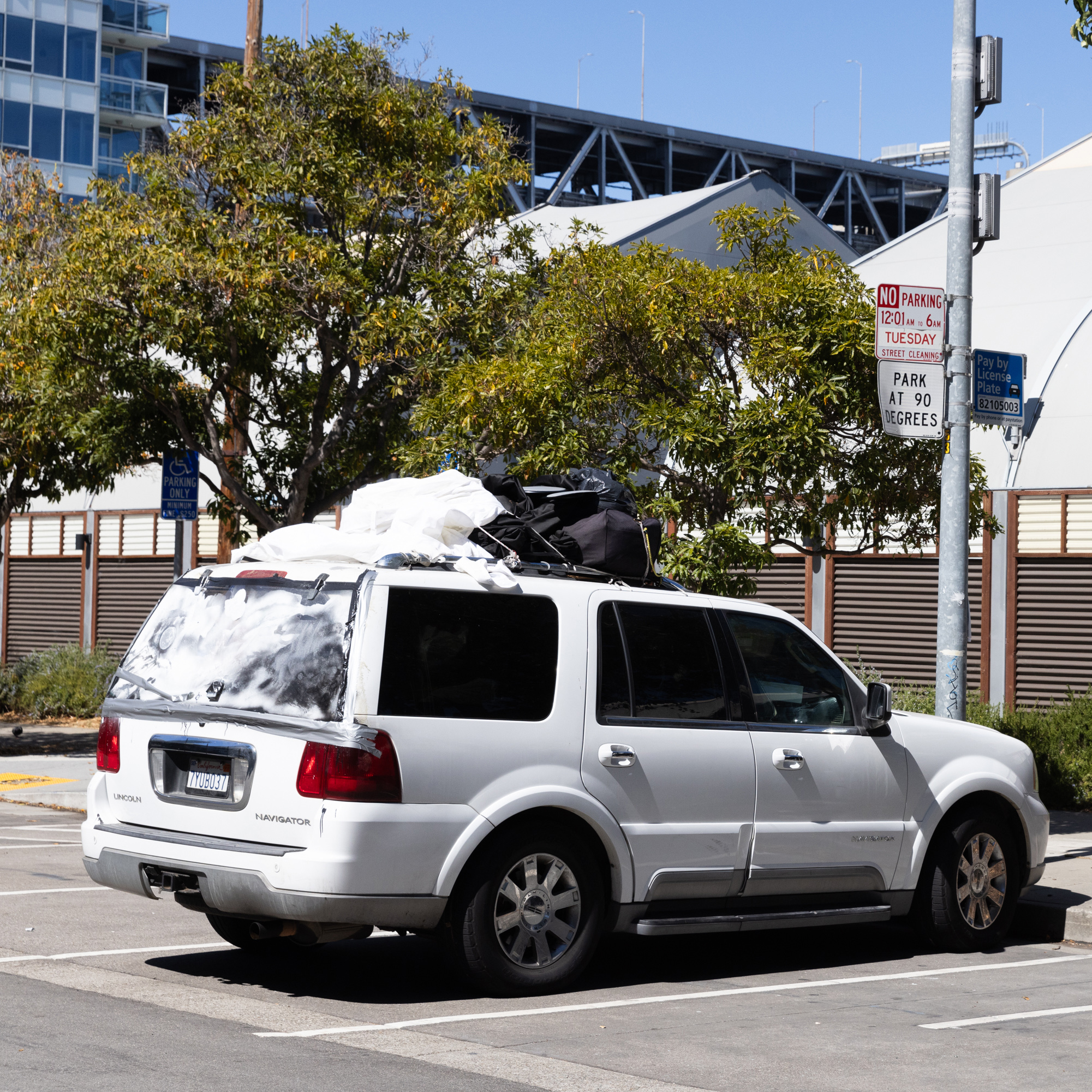 A white SUV with items tied on its roof is parked in a lot. Trees and signs, including a &quot;No Parking&quot; sign, are nearby, with buildings visible in the background.