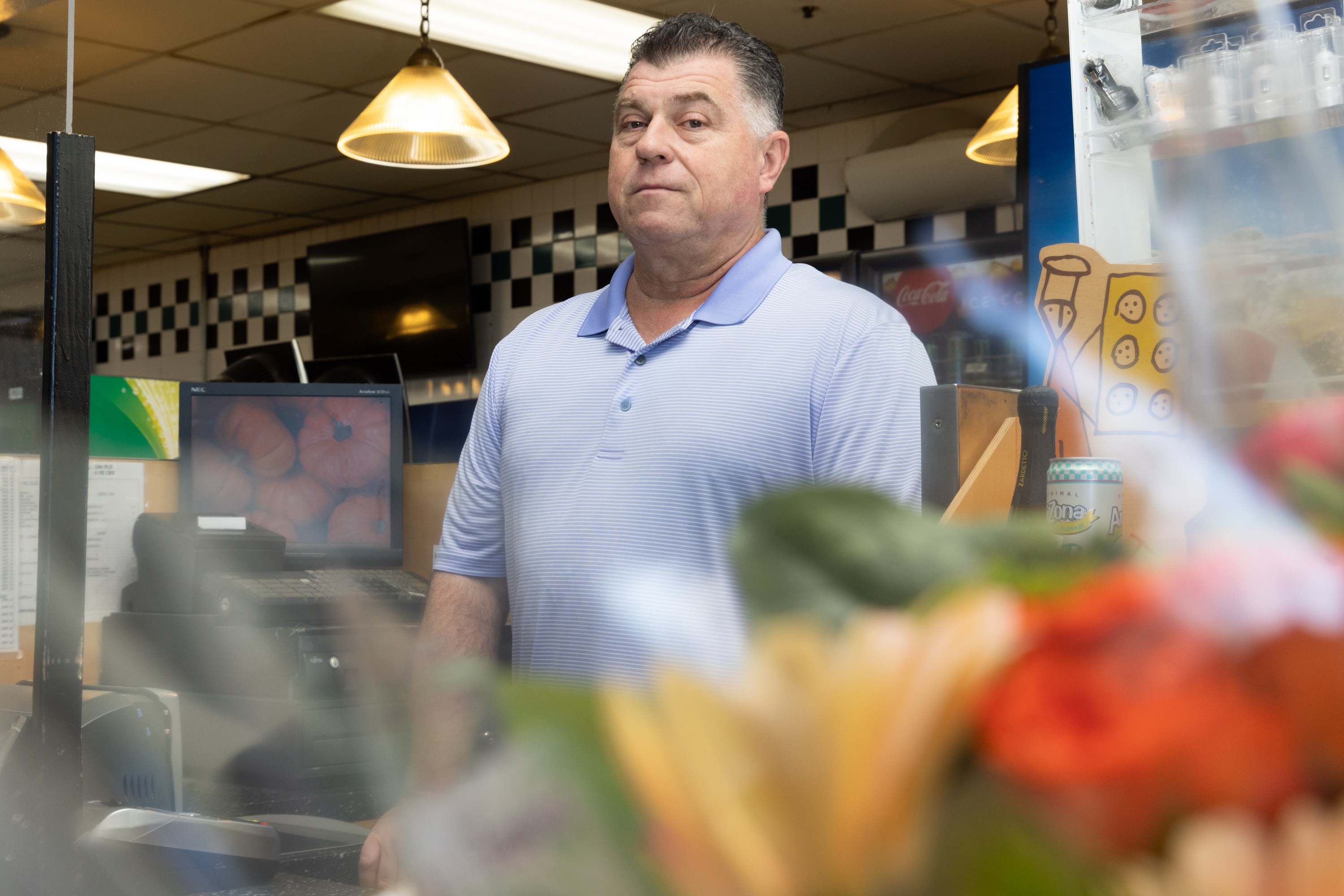 A man in a light blue polo stands behind a counter with a checkered wall. The foreground is blurred with colorful flowers. Hanging lights and a computer screen are visible.