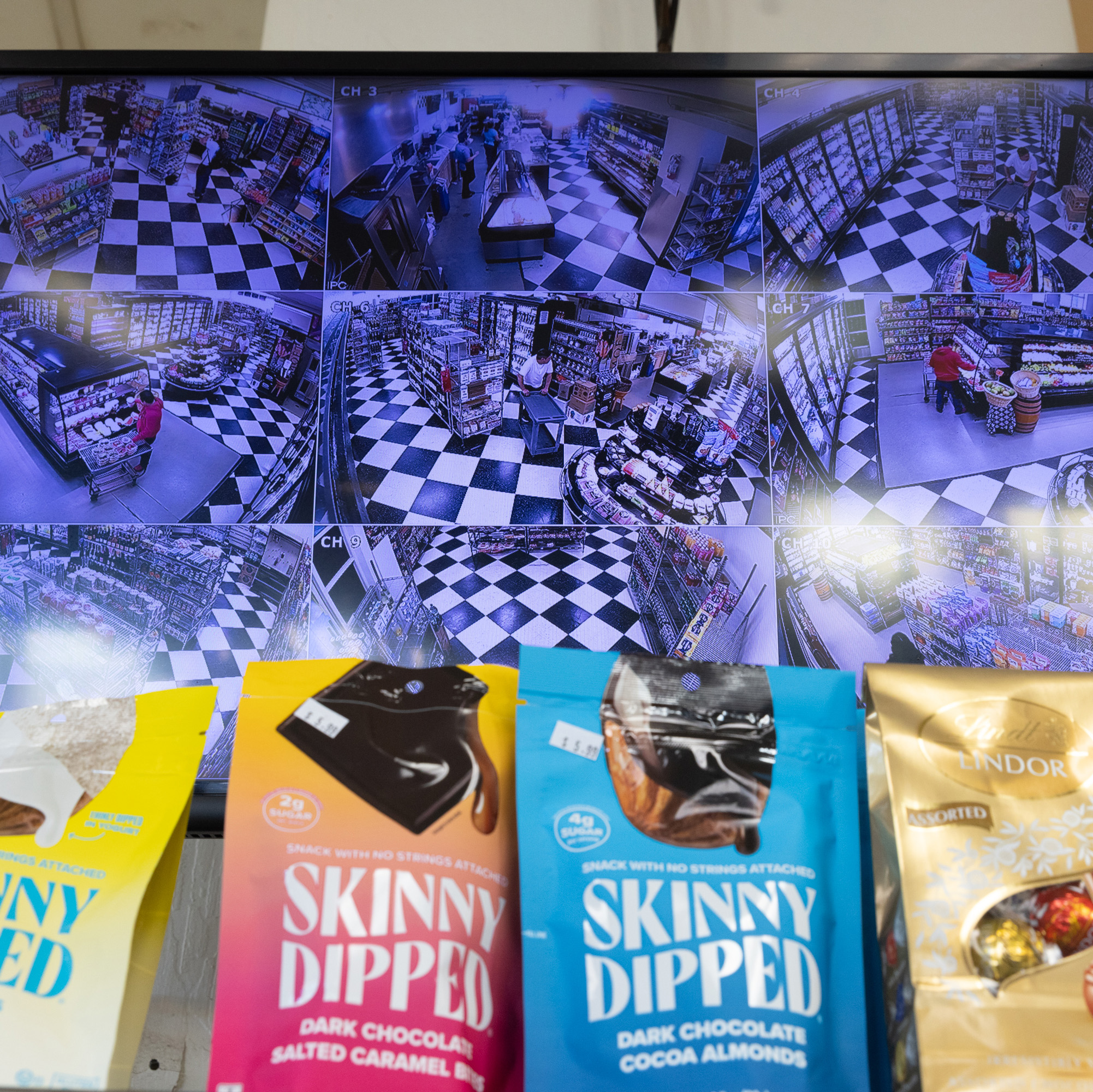 The image shows a security monitor with multiple camera feeds displaying a store with a checkered floor, and several Skinny Dipped snack bags are visible in the foreground.