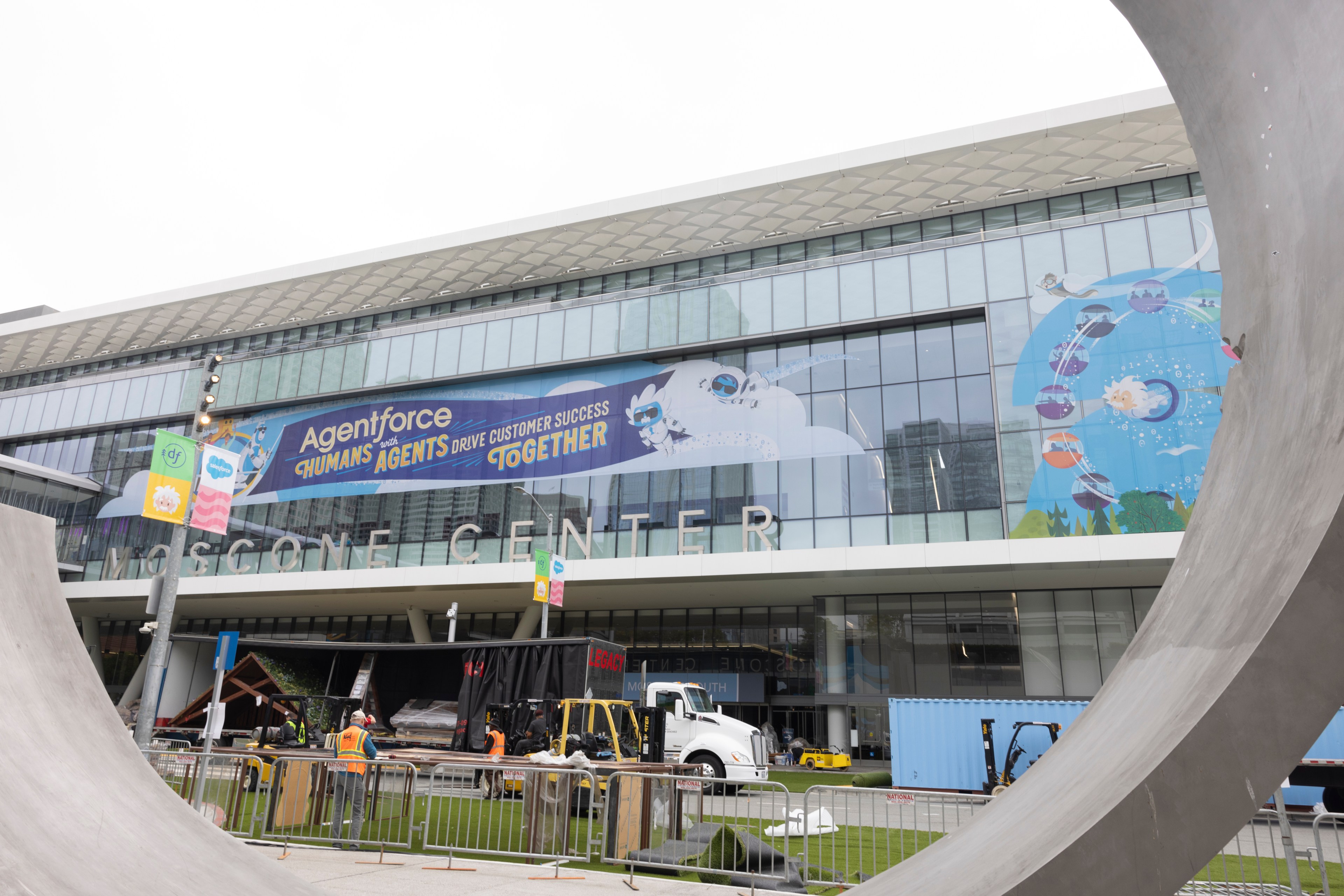 The image shows the Moscone Center with a large banner that reads, &quot;Agentforce with humans and agents drive customer success together.&quot; Workers are setting up outside.