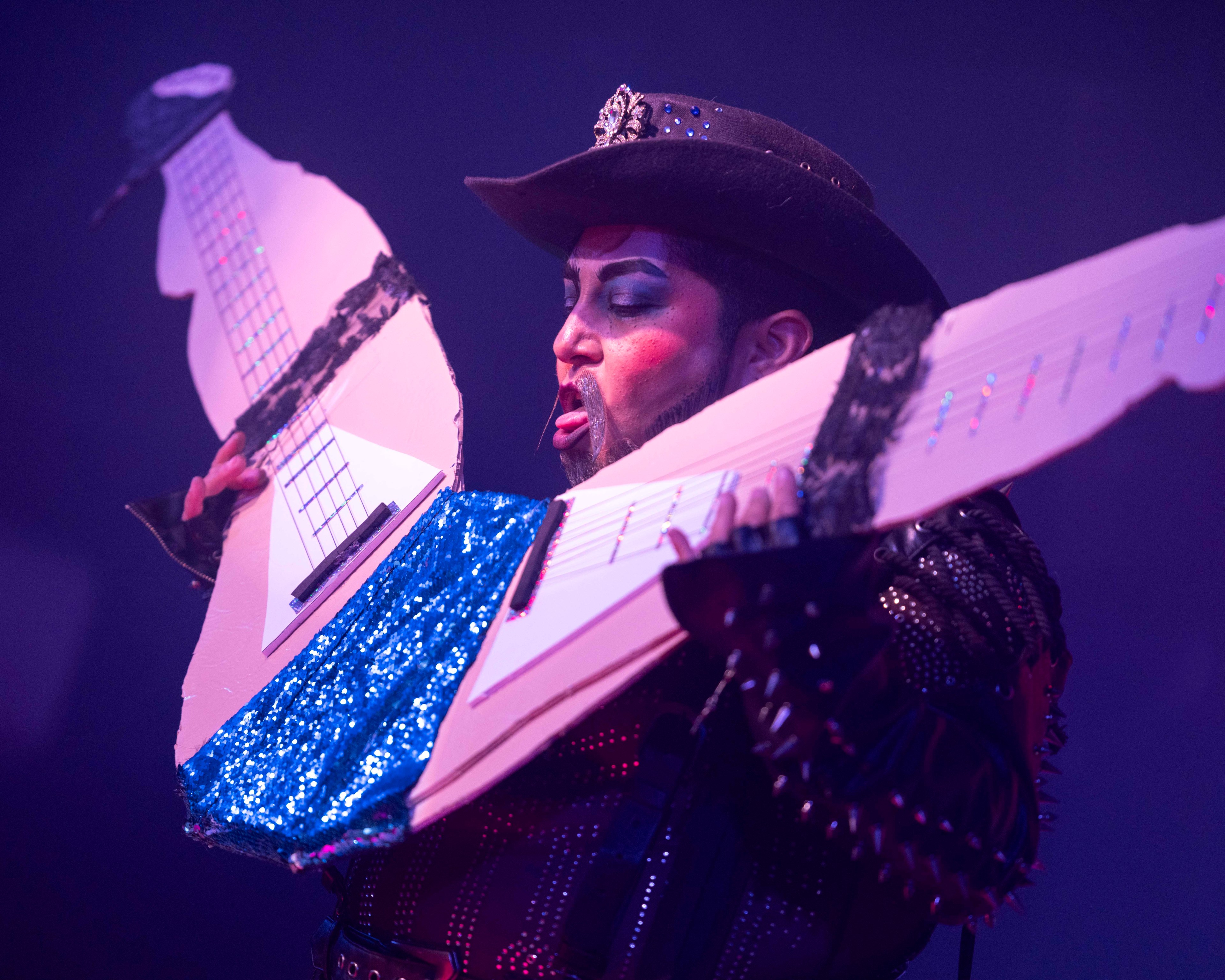 A performer in dramatic makeup and a studded outfit holds an oversized, double-neck cardboard guitar decorated with blue sequins, appearing mid-act on stage.