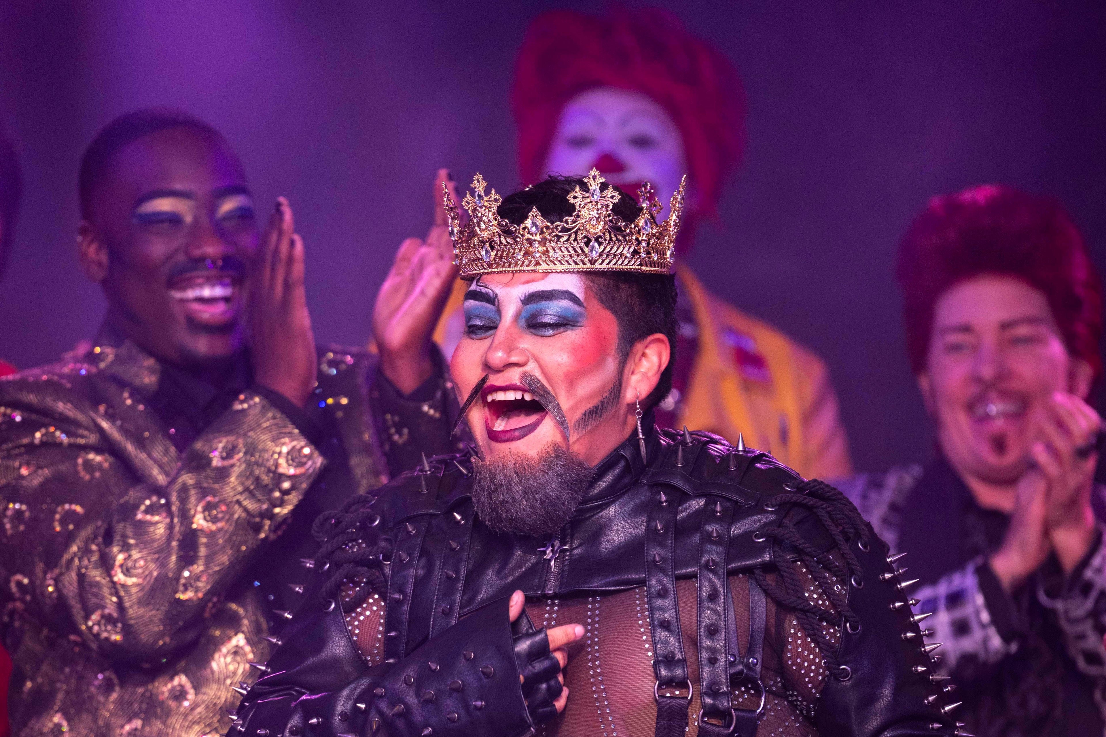 A person in a studded leather outfit and crown jubilantly holds a hat while being applauded by a diverse group of extravagantly dressed and makeup-clad individuals.