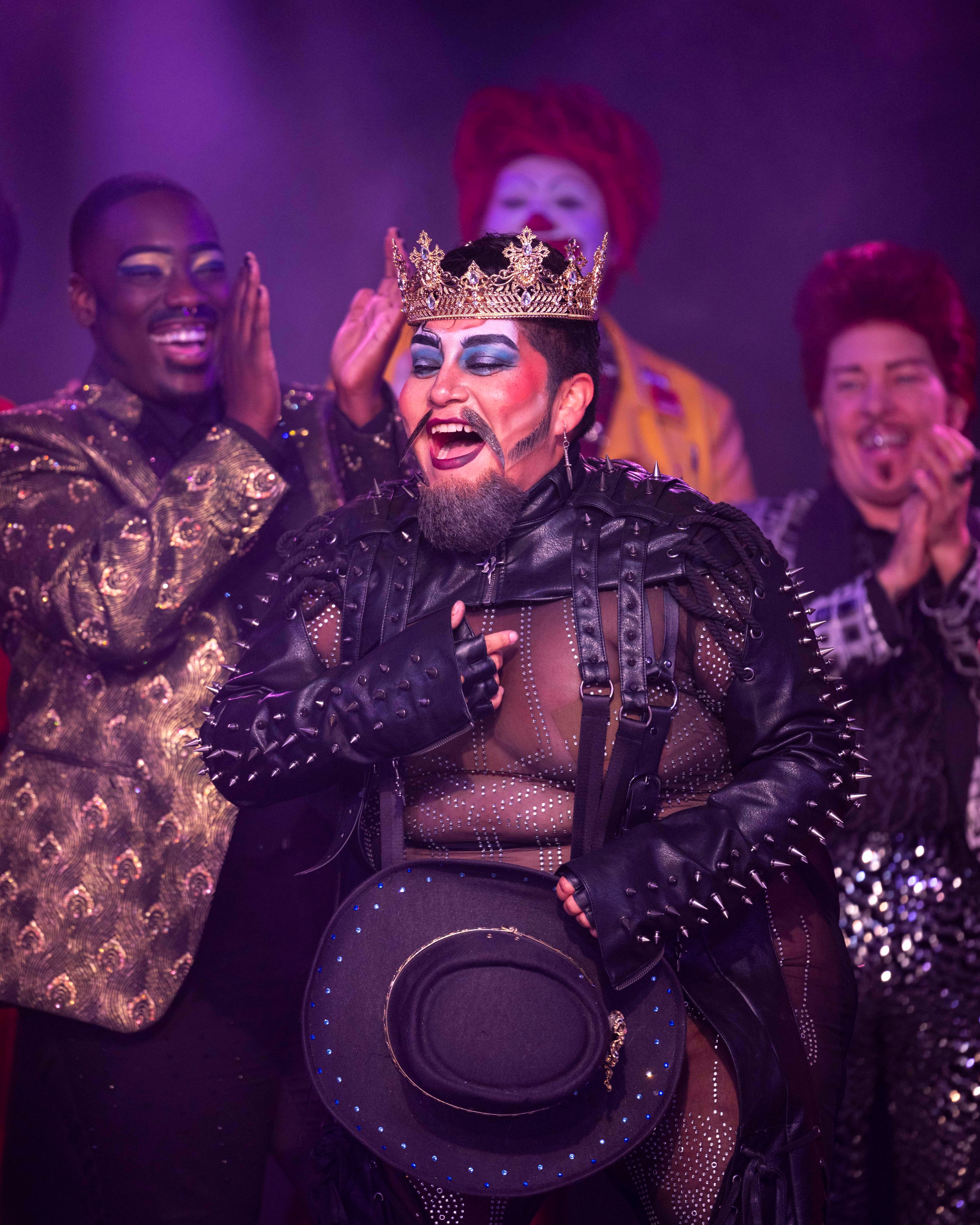 A person in a studded leather outfit and crown jubilantly holds a hat while being applauded by a diverse group of extravagantly dressed and makeup-clad individuals.