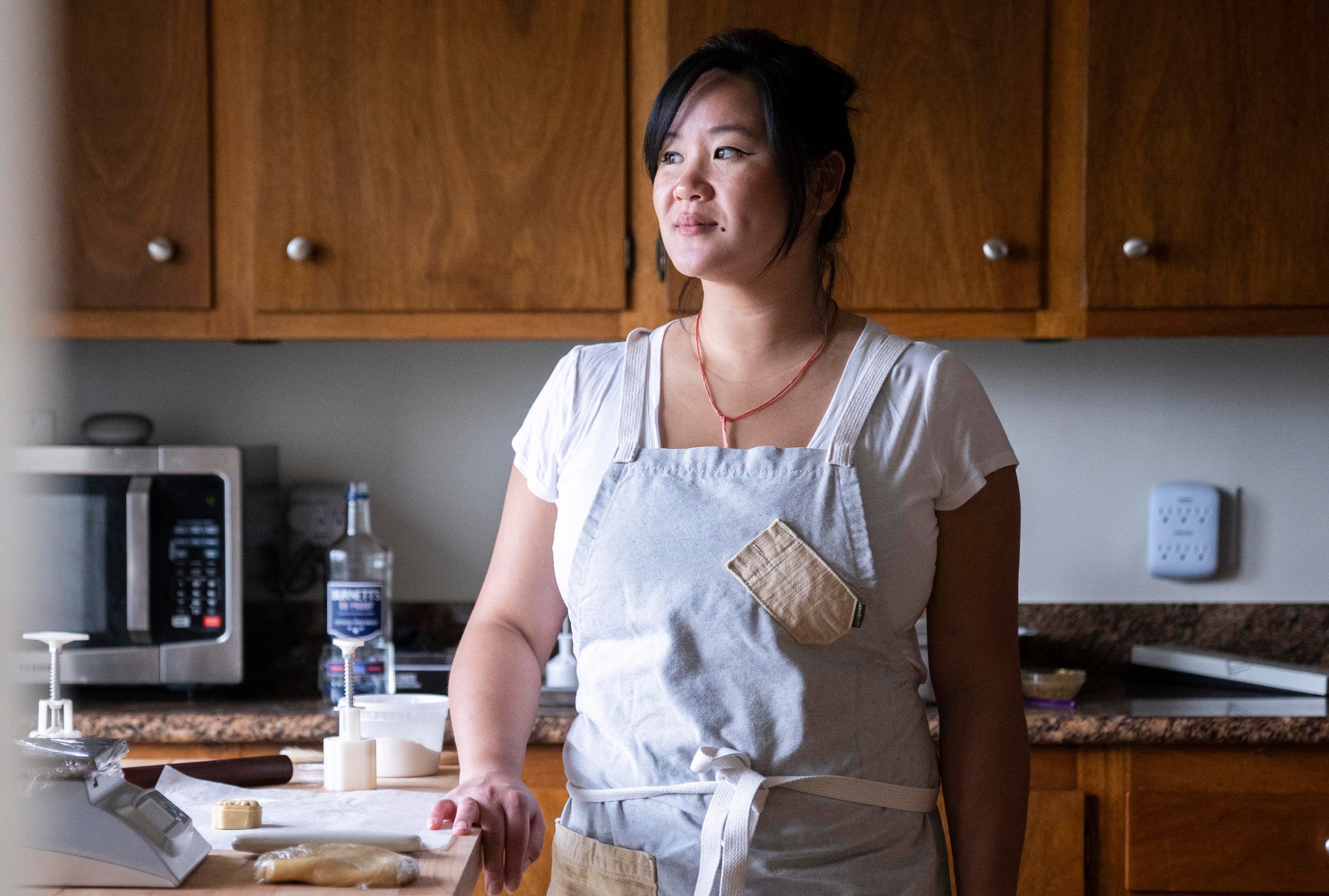 A person wearing a white shirt and light gray apron stands in a kitchen. Behind them, a microwave, wooden cabinets, a bottle, and cooking ingredients are visible.